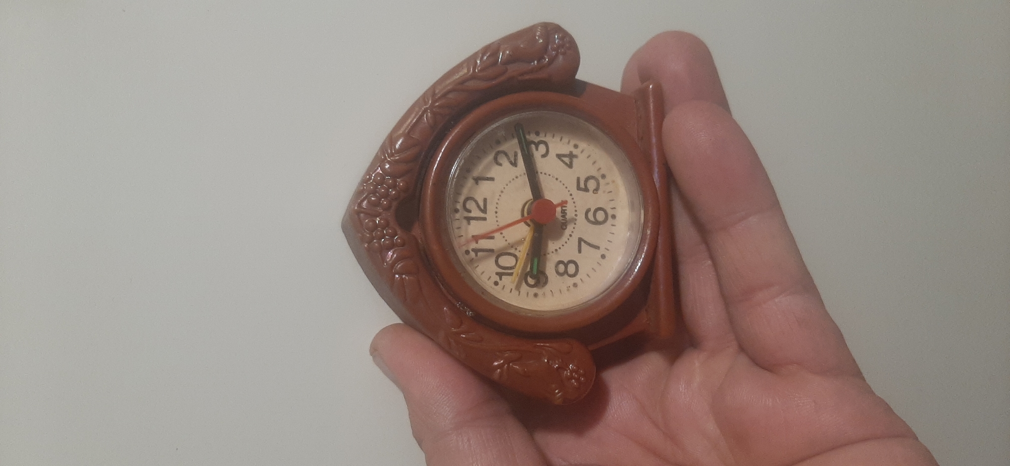 The image shows a close-up of a person's hand holding a small, brown, decorative alarm clock. The clock has a round face with white numbering from 1 to 12. The hands of the clock are visible: a red hour hand, a yellow minute hand, and a thin green second hand. The clock itself is encased in a brown, sculpted frame that appears to be made of resin, designed to look like ornate decorative elements, possibly vines or leaves. The hand holding the clock is a light-skinned adult hand. The background is a plain, light gray surface, suggesting an indoor setting. There is no clear indication of the time of day, weather, or specific location beyond the provided context of Yessentukskaya, Russia. The overall impression is that of a person showcasing or examining a small trinket. Visible text on the clock face reads "QUARTZ".