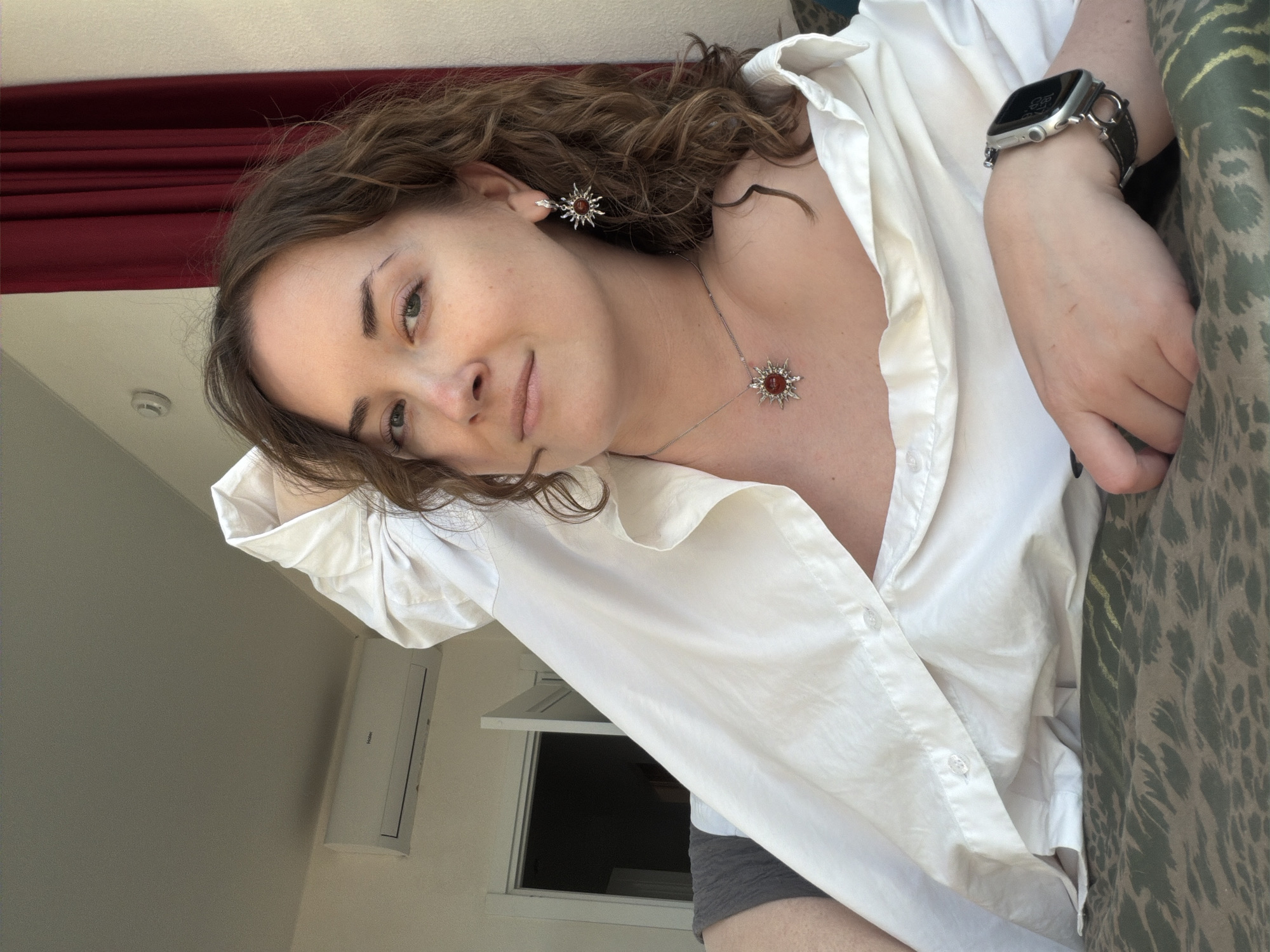 A portrait captures a woman with curly brown hair, adorned with starburst earrings and a matching necklace, lying horizontally with her head slightly tilted towards the viewer. She wears a loosely buttoned white shirt that reveals a portion of her chest. On her wrist, she sports a smartwatch with a black band. Behind her, there's a red curtain and a section of patterned fabric, and to her side, an air conditioning unit is mounted on the wall below a window. The location is unspecified.