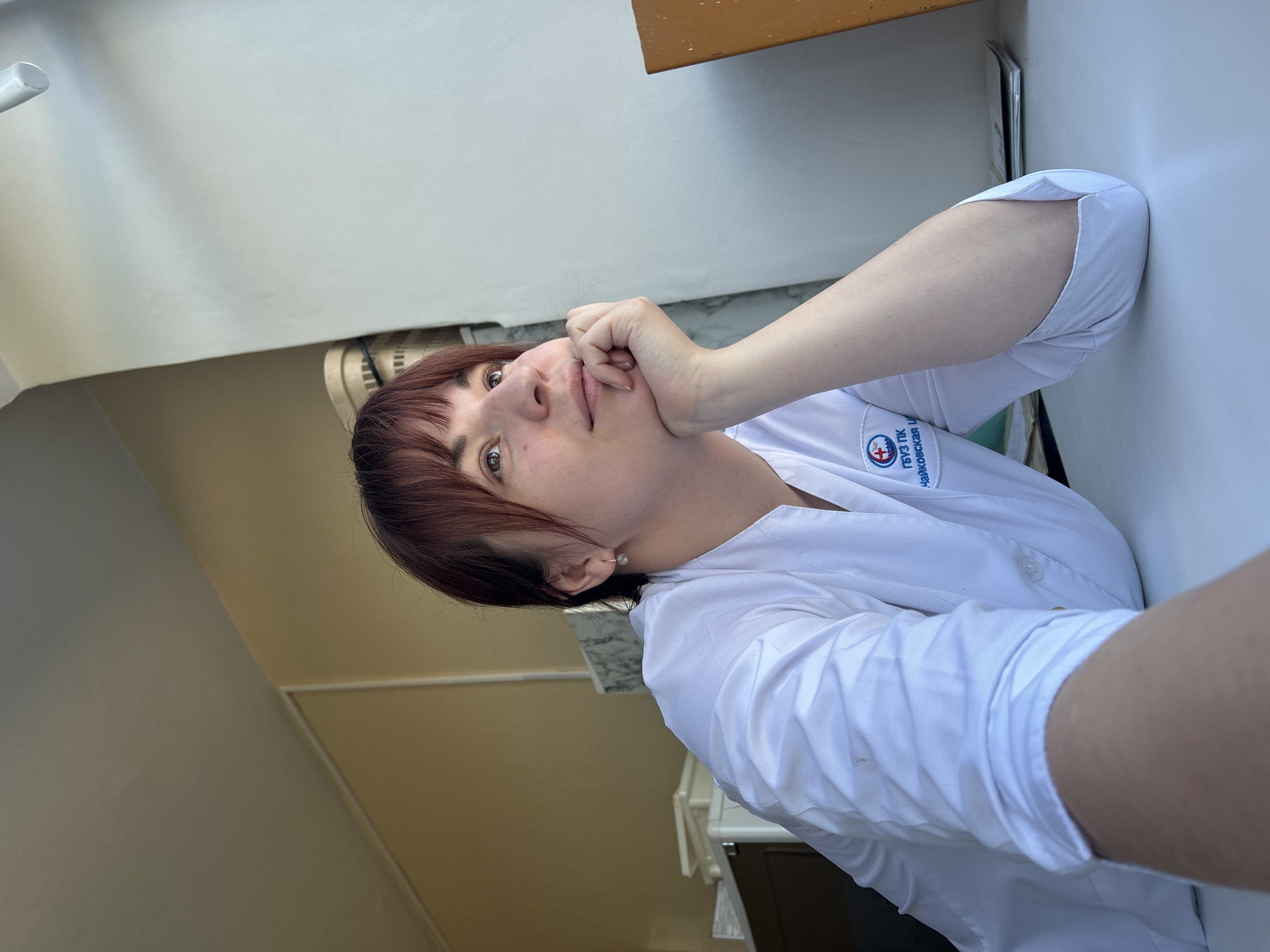 A woman with dark red hair is posing in a white medical coat. Her left hand is raised to her face, fingers near her lips. The location is Фоки, Russia, as indicated by the inscription on her coat which partially reads "...Чайковская И". The background is a combination of white walls and a partially visible cabinet with a wooden top. The lighting appears to be natural daylight.