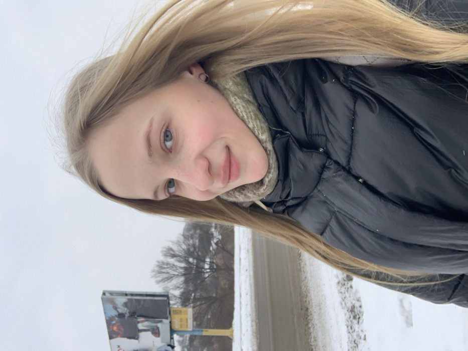 The image captures a young female outdoors in a winter setting in Bohuslav, Ukraine. The subject, positioned slightly right of center, presents a frontal view, displaying fair skin, blue eyes, and long blonde hair. She wears a black puffer jacket over a green/khaki knit neck warmer and a silver-colored hoop earring in her visible left ear. Her expression is a slight smile directed towards the viewer. In the background to the left, a large billboard mounted on a yellow and blue pole structure displays two advertisements; the left advertisement features a figure holding a torch. Below the billboard, a smaller yellow and blue sign with text and numbers is visible. A bare deciduous tree stands behind the billboard. The ground is covered with snow, and a dark road surface is visible in the lower-left quadrant. The sky is uniformly overcast and light grey.
