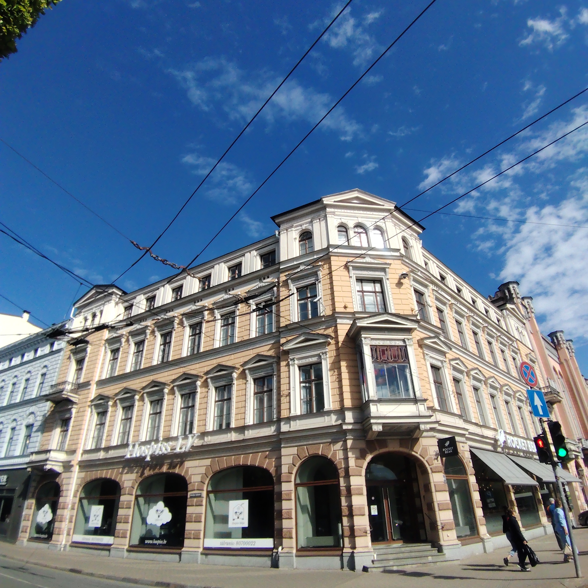 The image captures a street scene in Riga, Latvia, featuring a multi-story, pale yellow building with arched storefronts. The building houses at least two businesses, "Hospiss LV" and "Rocket Bean." Pedestrians are walking along the sidewalk. A traffic light shows green, and street signs are visible. Overhead, a network of power lines crisscrosses the clear, blue sky with scattered clouds. The architecture suggests a historical setting.