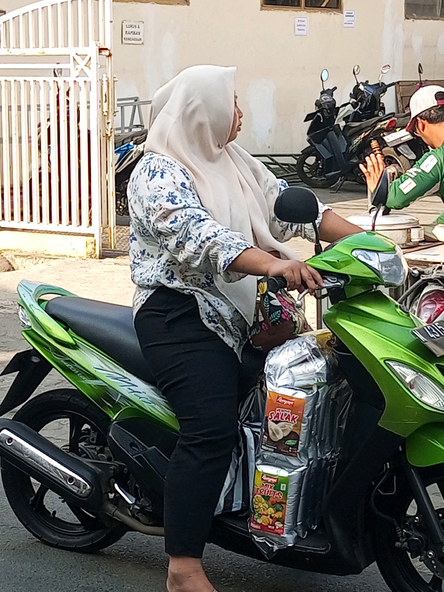 In Batu, Indonesia, a woman wearing a light-colored hijab and a floral-patterned blouse is seated on a green scooter. She is holding the handlebars with both hands, her left hand on the grip and her right hand operating the brake lever. Black pants cover her legs. Attached to the front of the scooter are two large, silver packages; the top package displays the word "SALAK" in red lettering, indicating it contains snake fruit. Below it, another package reads "MIX FRUITS". The scooter is parked on a paved surface, possibly a street or walkway, with a white metal gate visible in the background, along with several parked motorcycles and a building. The woman is looking to her right, off-camera.