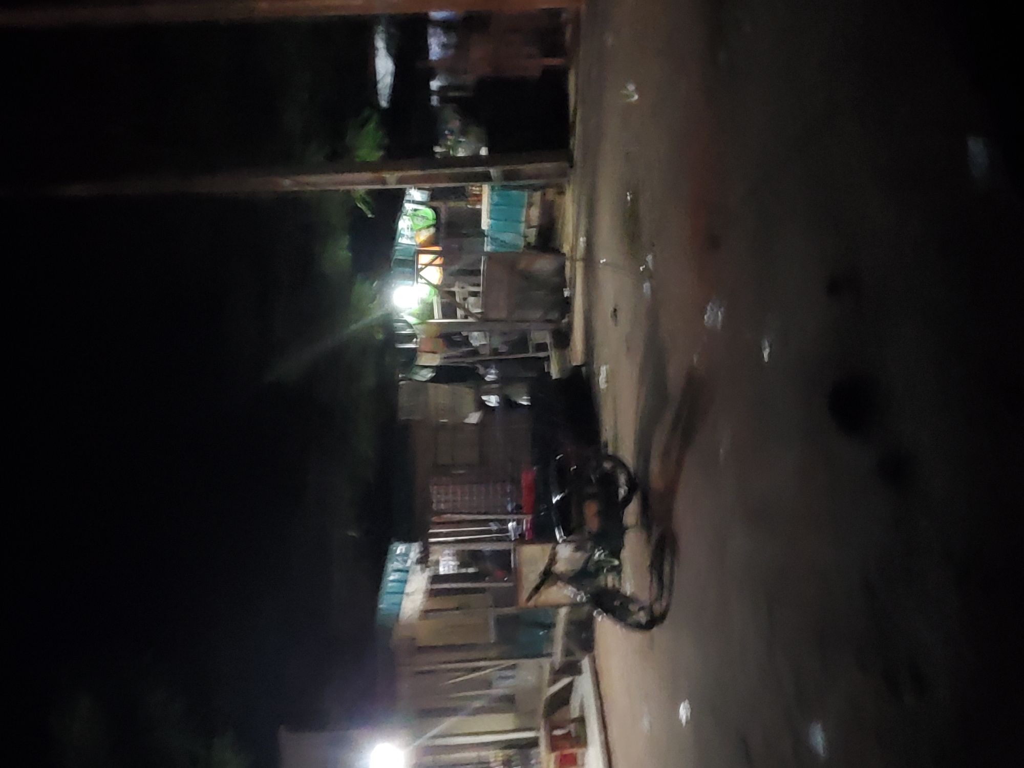 The image depicts a street scene at night in Jalingo, Nigeria. The primary subject is a parked motorcycle, partially visible on the right side of the frame. The scene appears to be a roadside stall or shop, with shelves and various items on display, though they are not clearly discernible due to the low light conditions. Structures with bright lighting, possibly stalls or buildings, are visible in the background, suggesting a market or commercial area. The ground is paved and appears wet, possibly from recent rain or cleaning, with scattered debris or reflections. The overall lighting is dim, with a few bright spots from artificial sources, indicating it is nighttime. There is no visible text in the image. The image is oriented vertically, and the quality is affected by blur and low resolution.