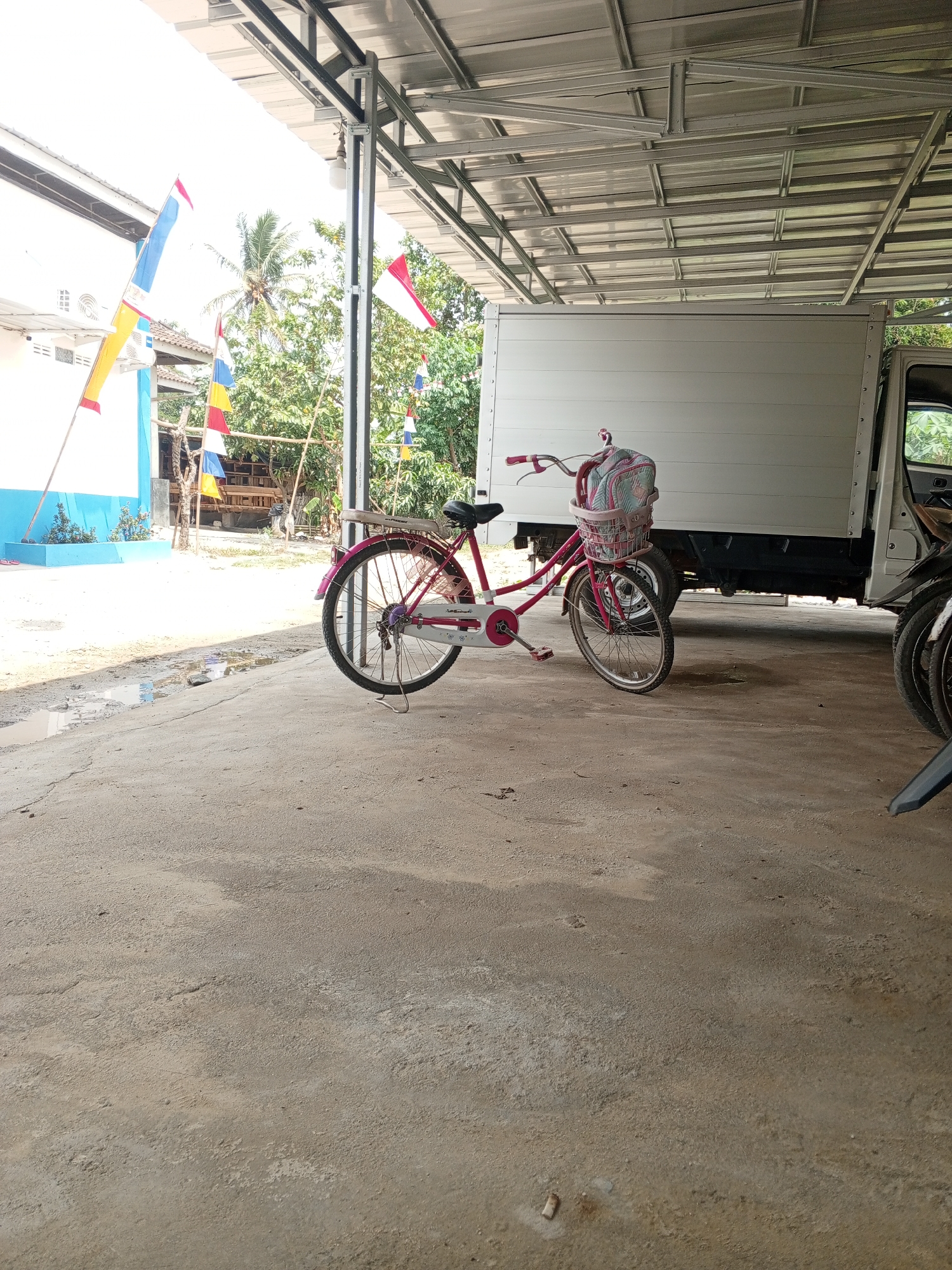 The image depicts an outdoor scene under a metal awning. In the foreground, a pink bicycle with a basket on the handlebars is parked on a concrete surface. The basket contains a pink and white patterned bag. Behind the bicycle is a white truck with its door open. The background reveals a building with blue and white accents, flags with red and white stripes, and lush green trees. The environment appears to be an open area, possibly a parking lot or a yard, in Sumber Rejo, Indonesia, indicated by the presence of Indonesian flags and tropical vegetation. The overall lighting suggests it is daytime.
