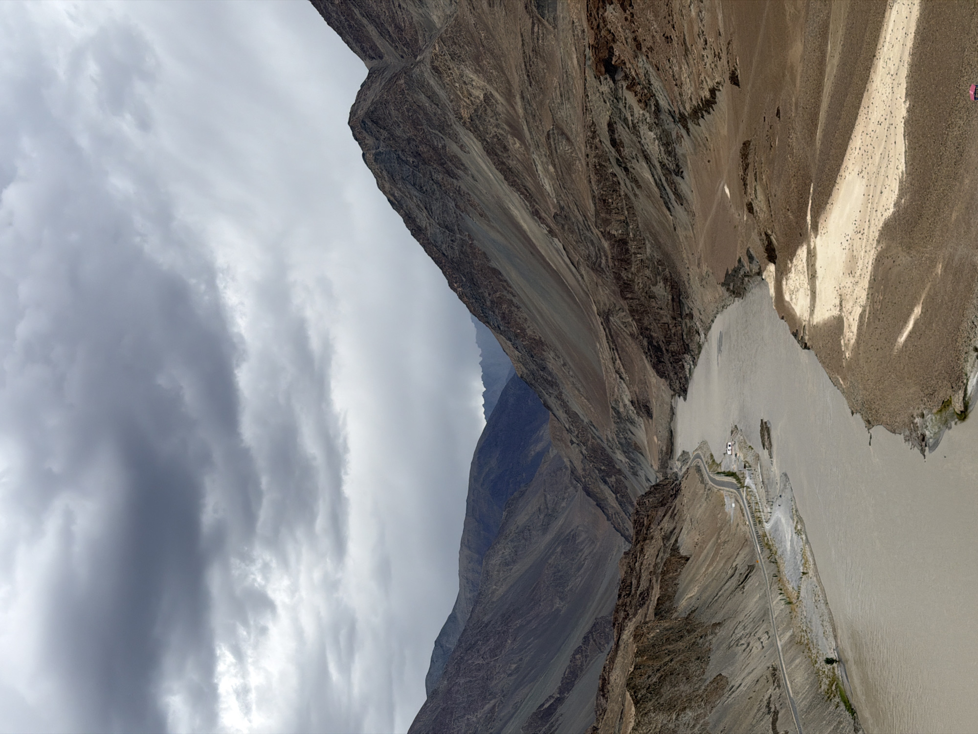 This image captures a wide, muddy river flowing through a dramatic mountain valley in Chumkar, India. The sky is overcast with heavy, grey clouds. The mountains on either side of the river are steep and barren, with exposed rock faces and sparse vegetation. A paved road winds along the left bank of the river, bordered by a small building and some scattered trees. The river itself is wide and slow-moving, with a light brown, sediment-rich color. The overall impression is one of rugged natural beauty and remoteness.