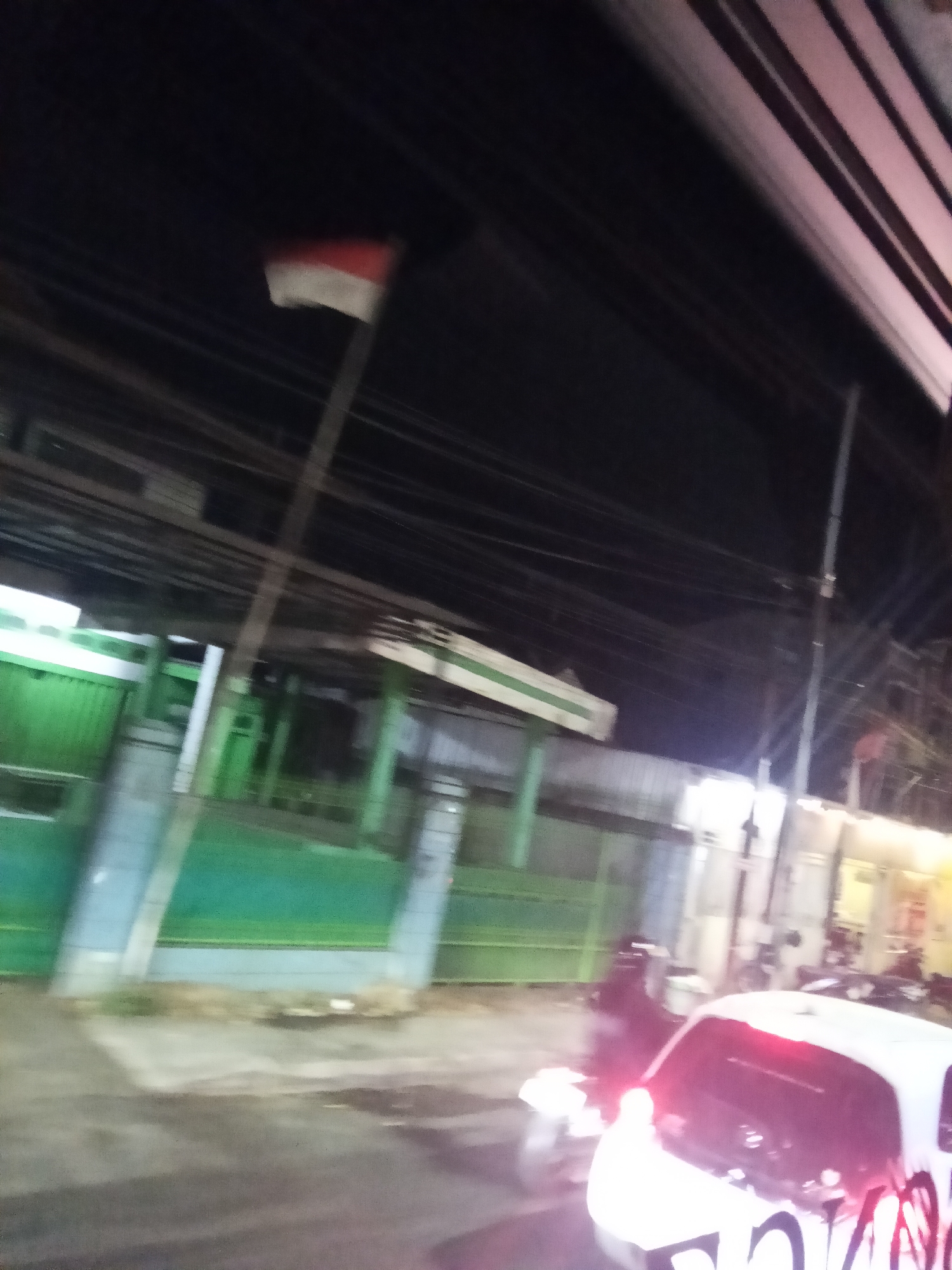 The image captures a dimly lit scene in Marumpa, Indonesia, likely at night. In the background, a red and white flag, possibly the Indonesian flag, flies from a pole against a dark sky filled with a dense network of overhead wires. To the left, a building with a green facade and white accents is visible, appearing to be a business or public facility. A street is in the foreground, with a white car parked or moving on it. The car's taillights are illuminated red, and the rear of the vehicle is covered in graffiti. Several indistinct figures are present near the car, and some motorcycles are also discernible, suggesting a street scene with traffic and pedestrian activity. The overall impression is that of a typical urban environment at night, with elements of daily life taking place under artificial and natural light.