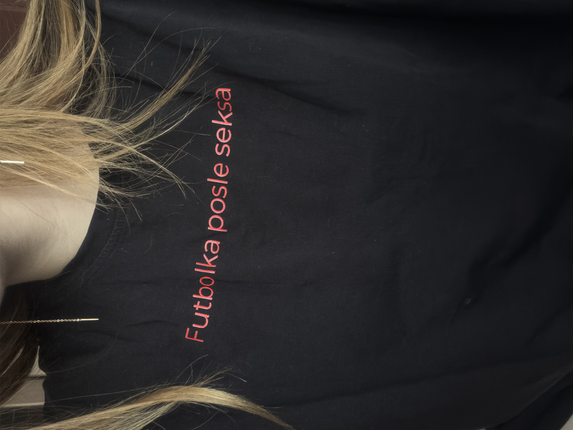 A photograph depicts a person wearing a black t-shirt with red text that reads "Futbolka posle seksa". The person has blond hair and is wearing a gold chain necklace. The background is dark and blurry, providing no additional context about the location or environment. No information to connect this image to the city of Chaikovsky, Russia.