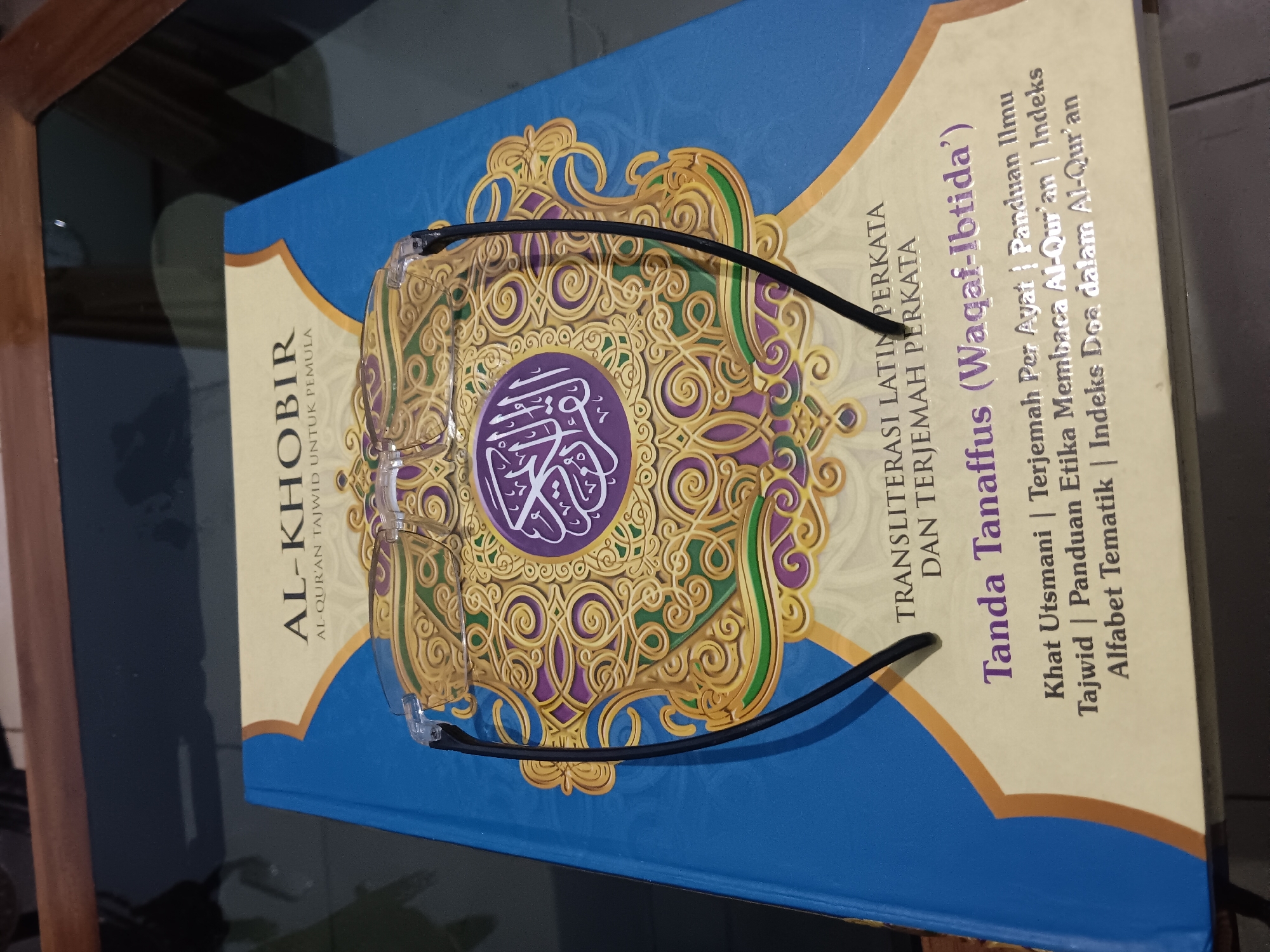 A clear face shield is placed on top of an open book with an Islamic design on its cover. The book has blue and yellow coloring with Arabic script. The face shield's clear plastic rests on the pages, with black arms extending outwards. The book is positioned on a dark, reflective surface, possibly a glass table, with wooden trim visible at the top of the frame. The scene is illuminated by ambient light, casting soft shadows.