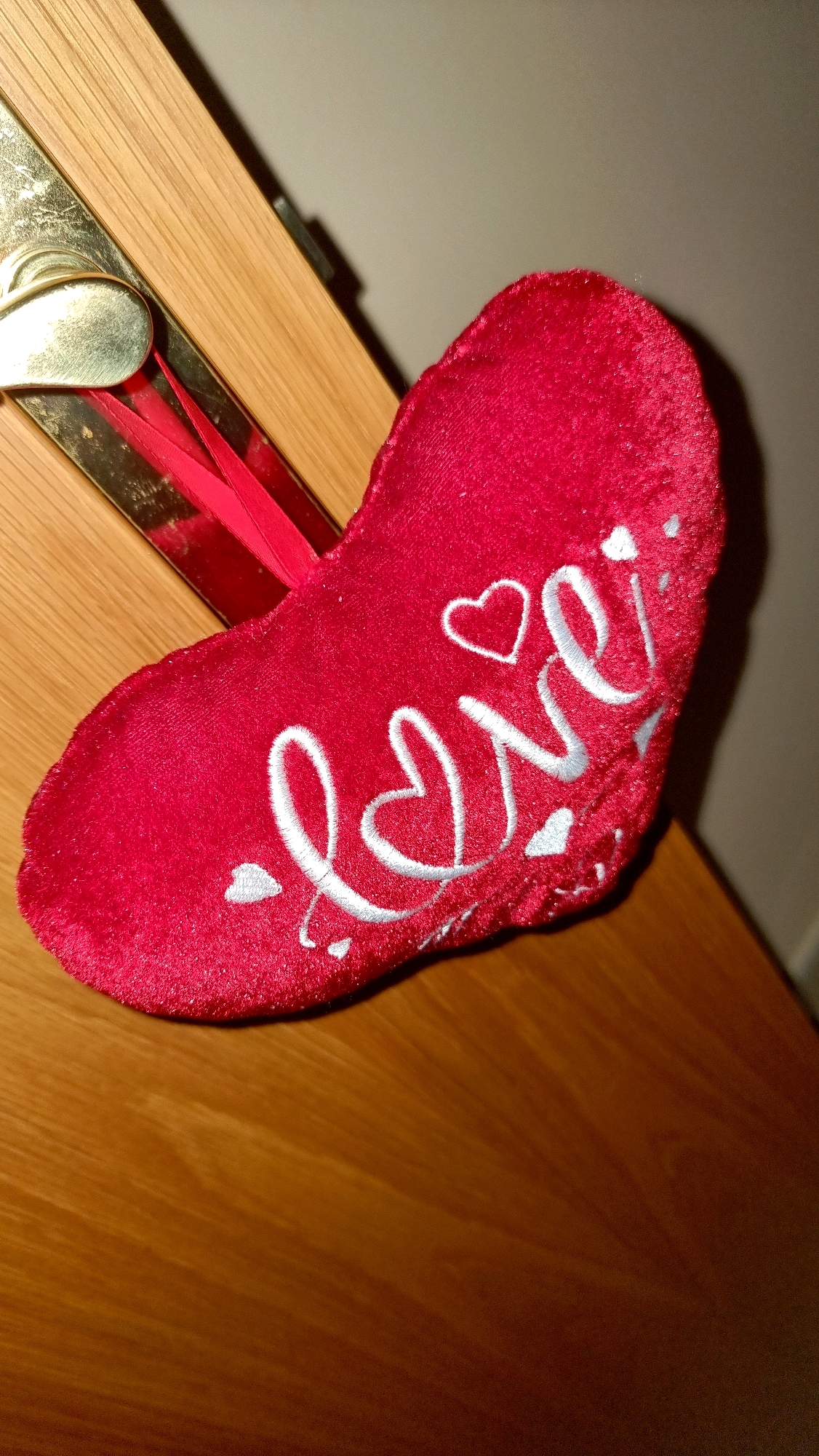A red heart-shaped ornament with the word "love" embroidered in white thread hangs from a wooden door. The ornament is plush and has a red ribbon attached. The door has a brass handle and appears to be made of oak. The background is a muted gray wall. This scene is located in Sant Joan Les Fonts, Spain.
