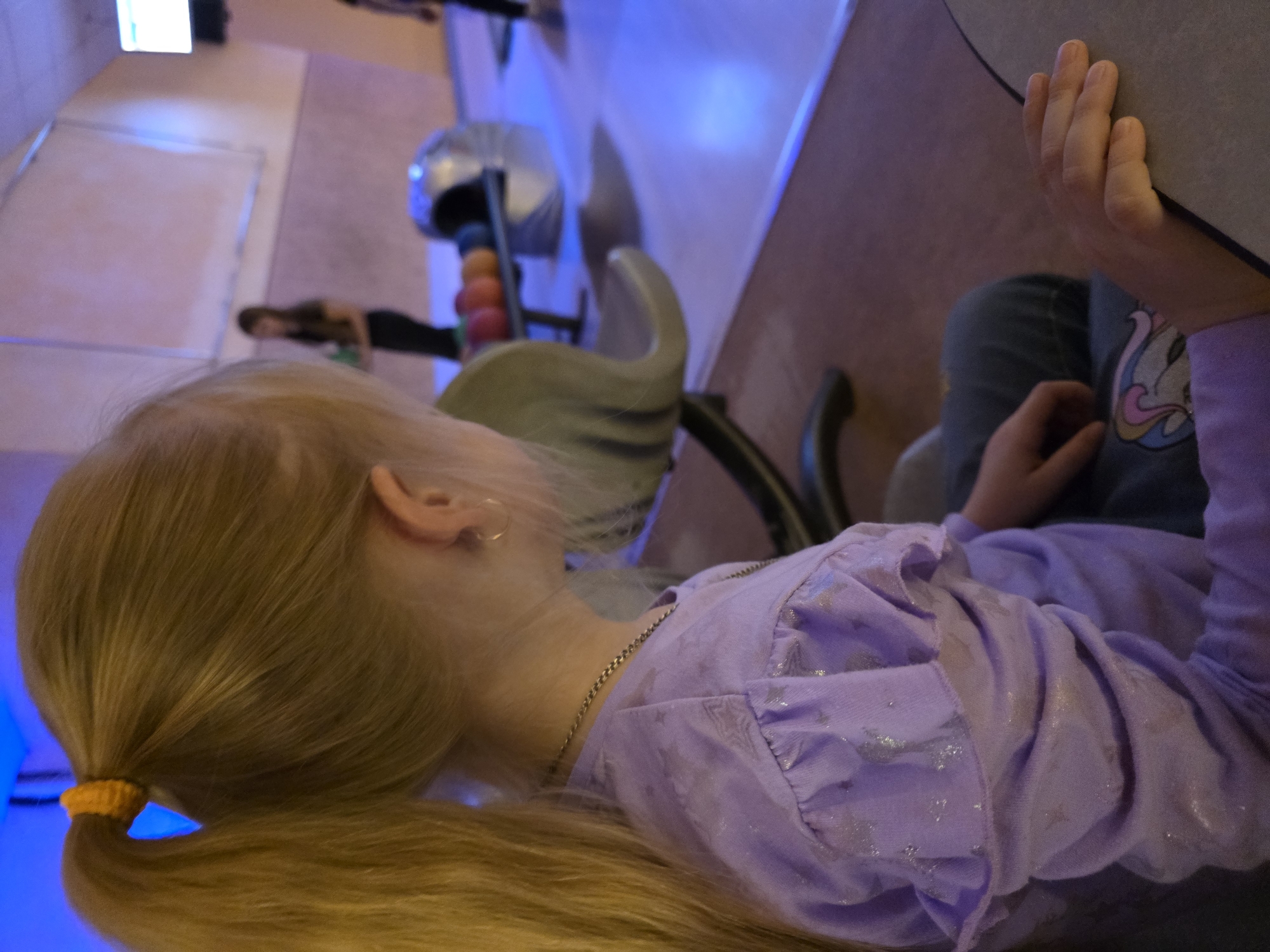 The image shows a blonde-haired girl wearing a purple shirt and jeans, sitting at a table. Her hair is pulled back in a ponytail with a yellow hair tie. She is at a bowling alley in Chaikovsky, Russia. Other people can be seen further down the lanes. A ball rack with bowling balls is visible. The lighting is somewhat dim with a bluish hue. The scene is taken from above and tilted, showing the girl from behind.
