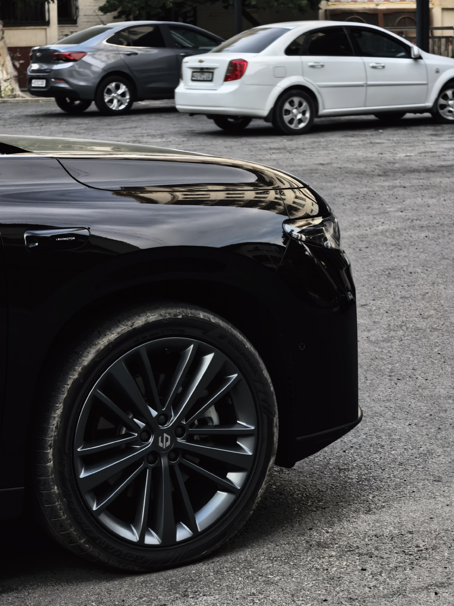 An eye-level, medium shot captures the front portion of a black car, focusing on its wheel and fender, with two other cars parked in the background. The black car's left front wheel, with a multi-spoke dark grey rim, is prominently displayed. The tire has visible tread and white lettering. To the left of the wheel, on the car's body, a black door handle with the inscription "LEAPMOTOR" is visible. The background features a grey sedan on the left and a white sedan on the right. The ground is paved with asphalt. The scene is outdoors, suggesting a city environment.