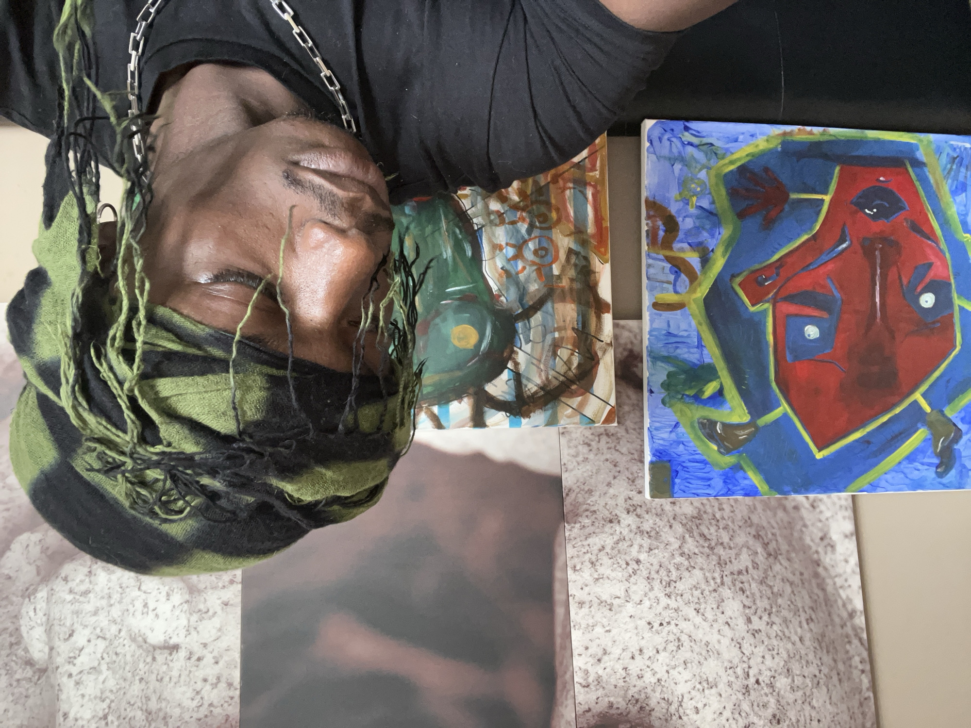 A man wearing a black shirt, green and black headscarf, and silver chain is lying down with his eyes closed. Next to him are two paintings on canvas. The painting on the right has a red figure outlined in yellow against a blue background. The painting on the left features green, yellow, and brown colors. The background is mottled and the location cannot be determined from the image provided. There is no clear evidence of the city of Barcelona, Spain.