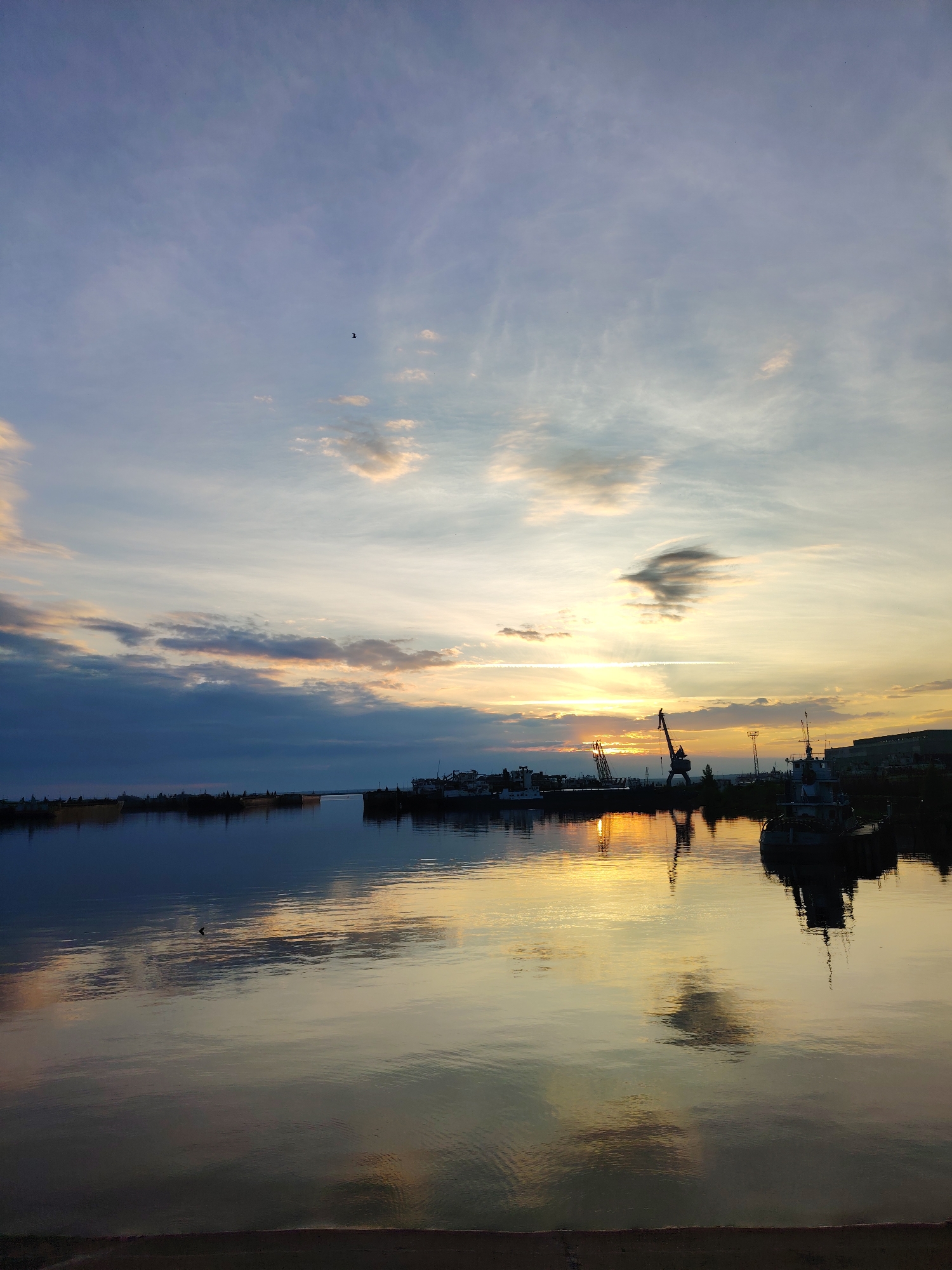 The image depicts a serene waterfront scene at sunset in the city of Chaikovsky, Russia. The sky is a canvas of soft blues and oranges, with wispy clouds catching the last rays of sunlight. The water in the foreground is calm and reflects the sky and the silhouettes of various vessels docked along the shore.
Several boats and ships are visible, including what appears to be a tugboat and larger cargo ships. Towering cranes stand in the background, hinting at the industrial nature of the port. The reflection of the setting sun on the water creates a warm, golden glow, adding to the tranquil atmosphere. The overall impression is one of peacefulness and the quiet end of a day by the water.