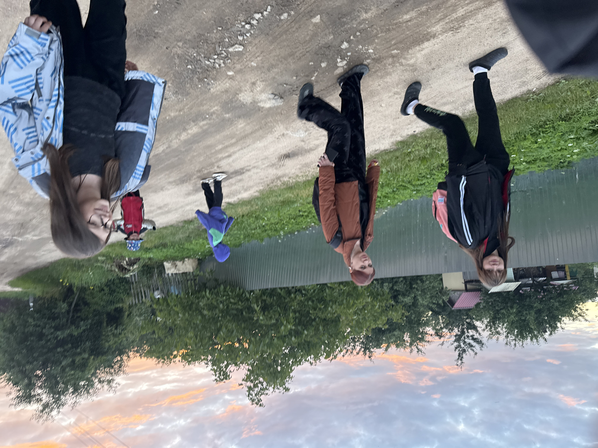 In Desnogorsk, Russia, at dusk, a group of five individuals, four visibly and one partially, traverse a dirt road bordered by lush green vegetation and a corrugated metal fence. The sky above is a canvas of soft, diffused light, with streaks of pink and orange clouds indicating the setting sun.
In the foreground and to the right, a young girl with long brown hair and glasses is captured from the chest up. She wears a patterned blue and white jacket over a dark top and black pants, her head turned to her right as if engaging with someone off-camera. Her expression appears pleasant.
To her left, further down the road, a person with brown hair and wearing a brown jacket over black pants with a star pattern walks away from the viewer. They carry a dark backpack. Their head is turned slightly to their left.
To the left of the person in the brown jacket and slightly ahead, a young woman with long brown hair walks briskly. She is dressed in a black jacket with white stripes, black pants, and black slip-on shoes. A pink backpack is visible on her shoulders. Her gaze is directed forward.
Further up the road, a child in a blue hooded jacket and purple pants runs ahead. Another child, wearing a red jacket and a patterned blue hat, stands facing the left side of the road.
The environment suggests a rural or suburban setting on the outskirts of Desnogorsk, with simple houses visible behind the fence in the background, and trees lining the right side of the road. The lighting conditions indicate it is late evening, with the sky still holding some color from the day.