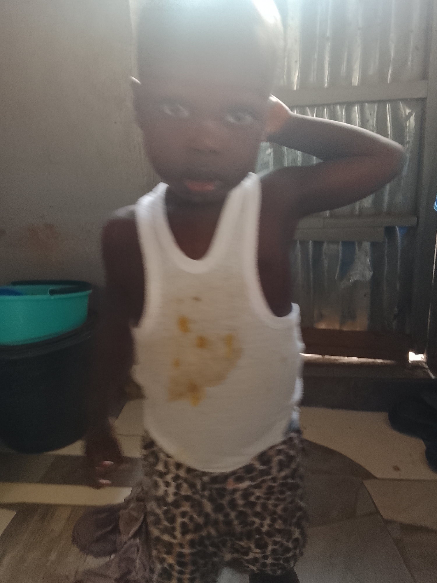 A young child stands indoors. The child is wearing a white tank top with a few visible stains on the front and animal-print pants. The child's right hand is raised and touching the right side of their head. Behind the child, there is a corrugated metal wall. To the left of the child, there is a black container with a blue bucket inside. The floor is tiled. The location is indeterminate, but stated as Abuja, Nigeria.