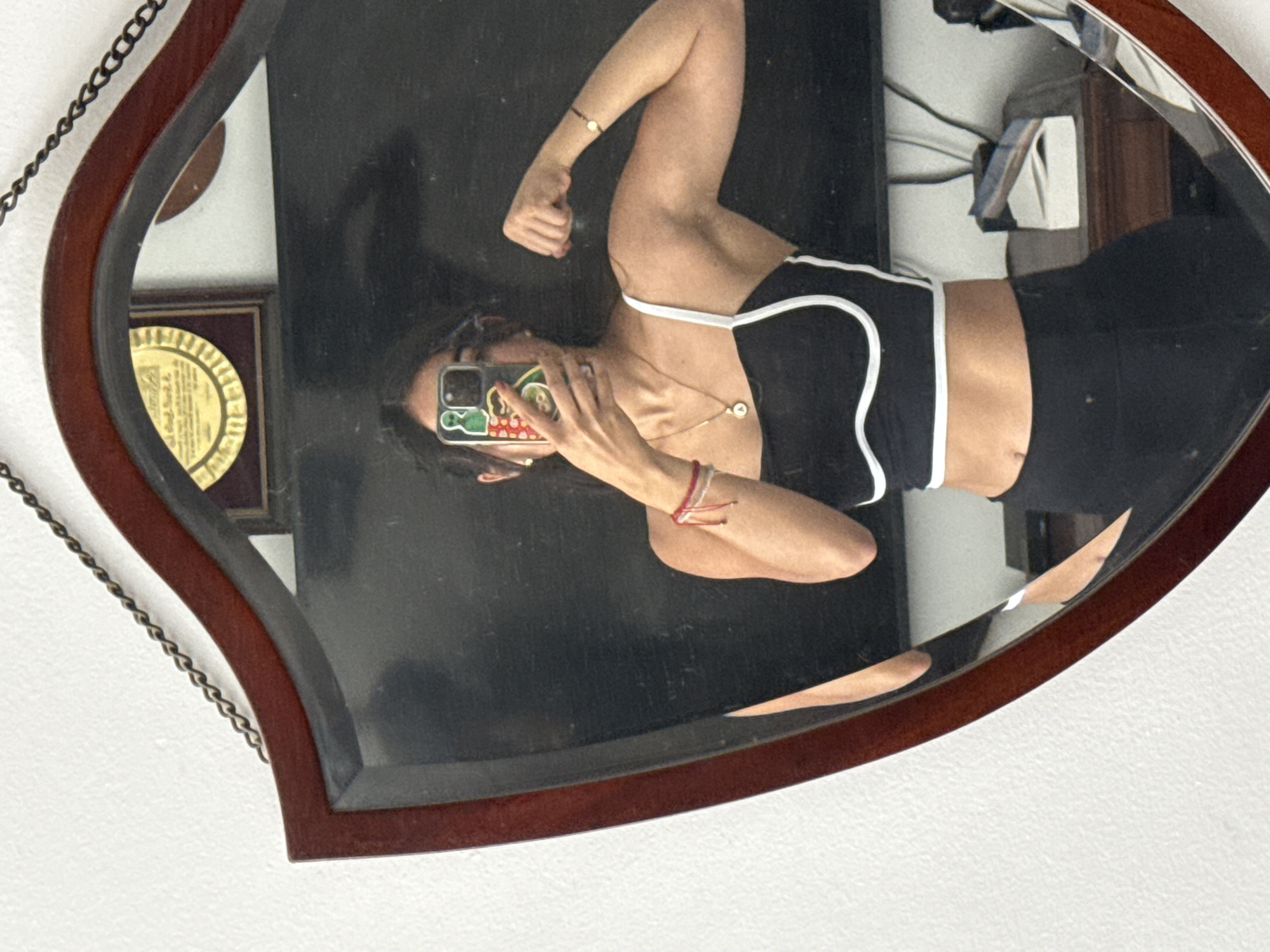 A person is reflected in a brown, leaf-shaped mirror hanging on a white wall. The person is holding a cell phone, obscuring their face. They wear a black top with white trim, black pants, and several bracelets. The background includes a dark door and framed certificates or awards. The image was most likely taken indoors. The location of the city of Puebla, Mexico, cannot be determined from the image.