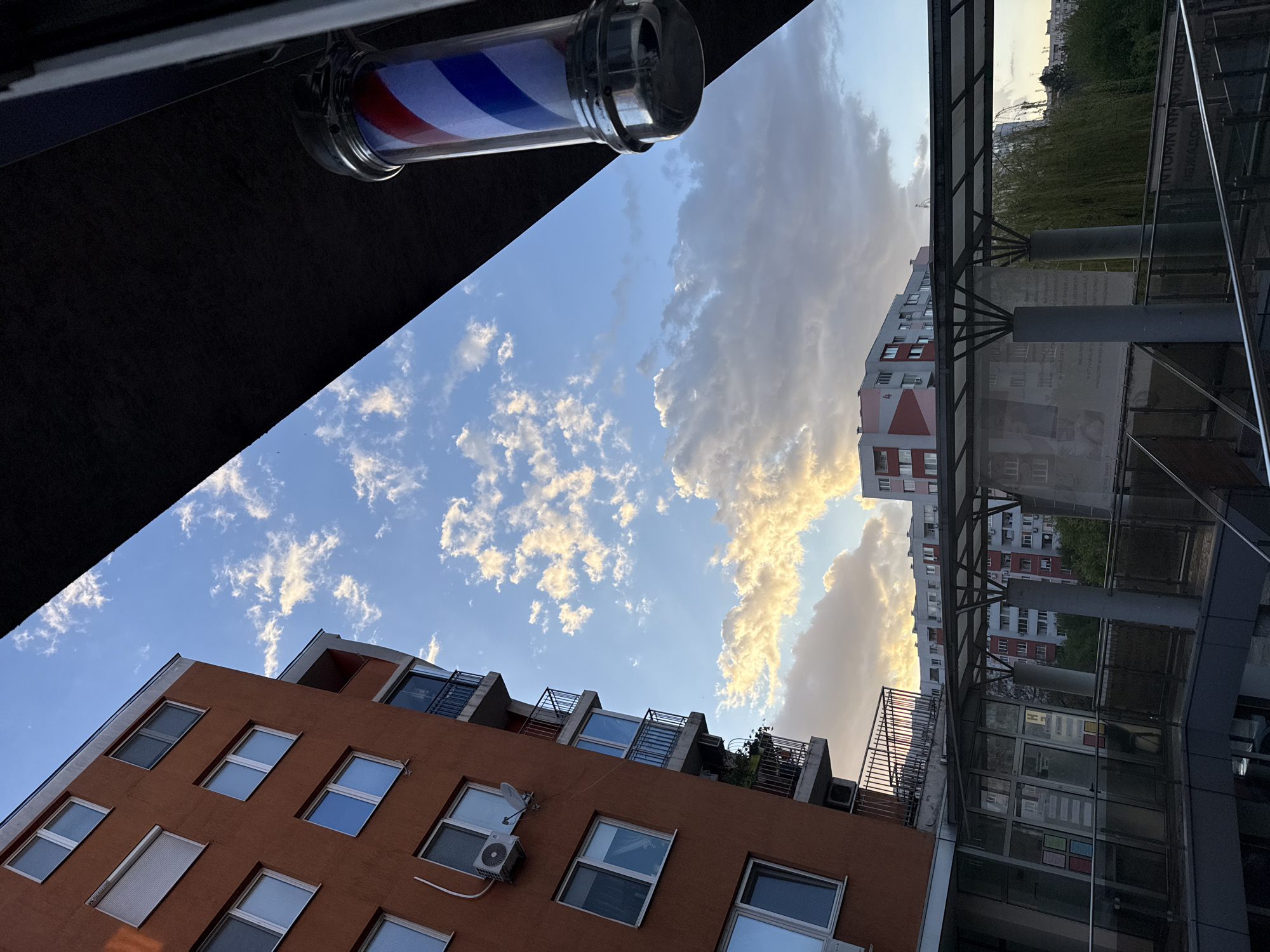 The image, taken from a low angle, features a barbershop pole in the upper left corner, characterized by red, white, and blue stripes. The view extends upward to reveal a sky with scattered clouds, exhibiting a gradient of blue and golden hues. Buildings are visible in the foreground and midground. A brick-colored building with multiple windows and small balconies is situated on the left. A taller, gray and red building stands in the background. A modern structure with glass and metal framing appears on the right, possibly a pedestrian walkway or part of a commercial building. Given the architectural style and details of the urban environment, the location is likely in Sofia, Bulgaria. There are no people visible in the frame.