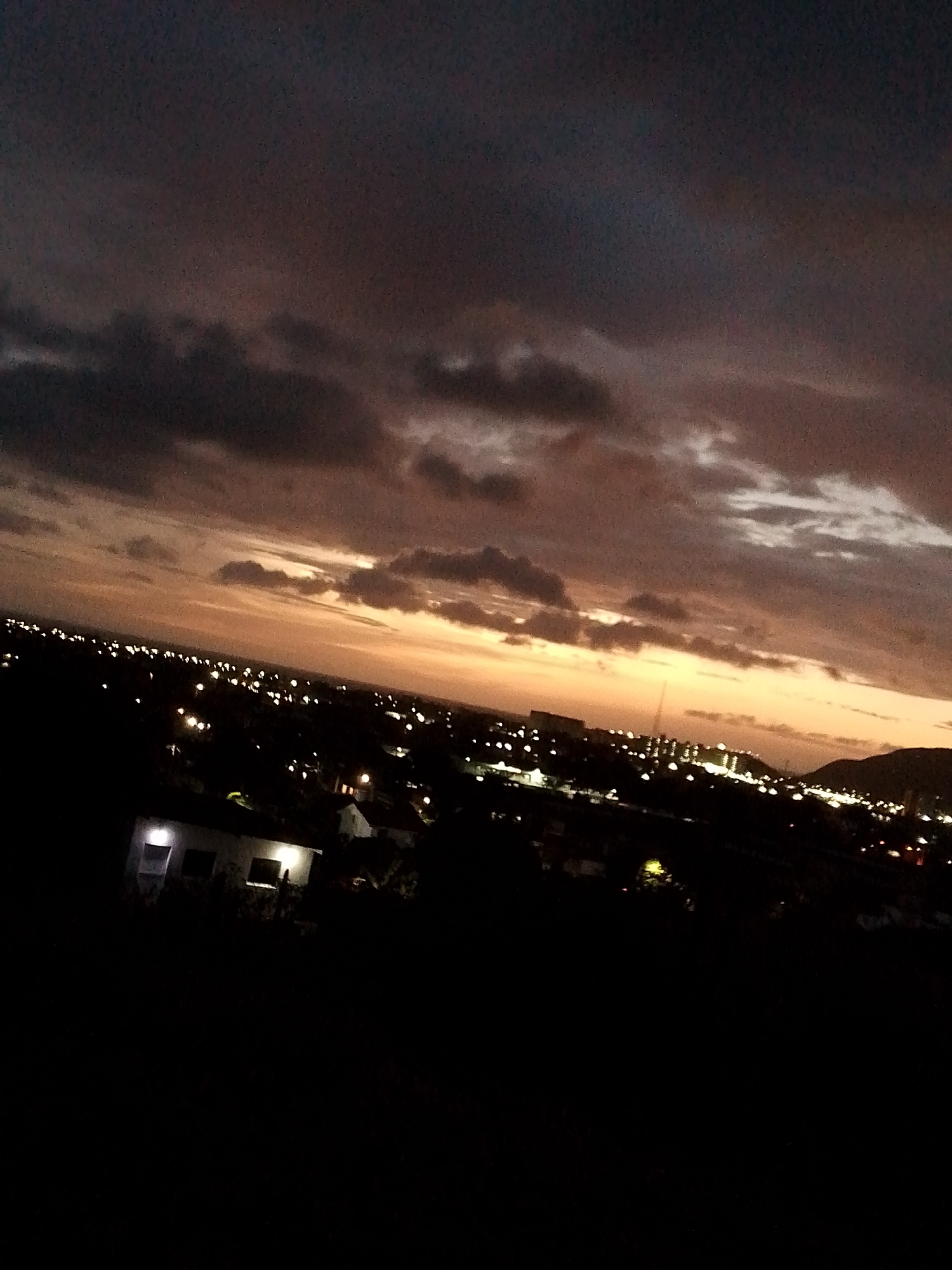 The image presents a nighttime cityscape from Altagracia, Venezuela. The sky is dominated by dark, layered clouds tinted with shades of orange and purple, indicative of a sunset or twilight. Below the horizon, numerous lights from buildings and streetlights illuminate the city. A structure with lit windows is visible in the foreground, while darker areas suggest foliage or unlit parts of the landscape. A mountain silhouette appears on the right horizon.