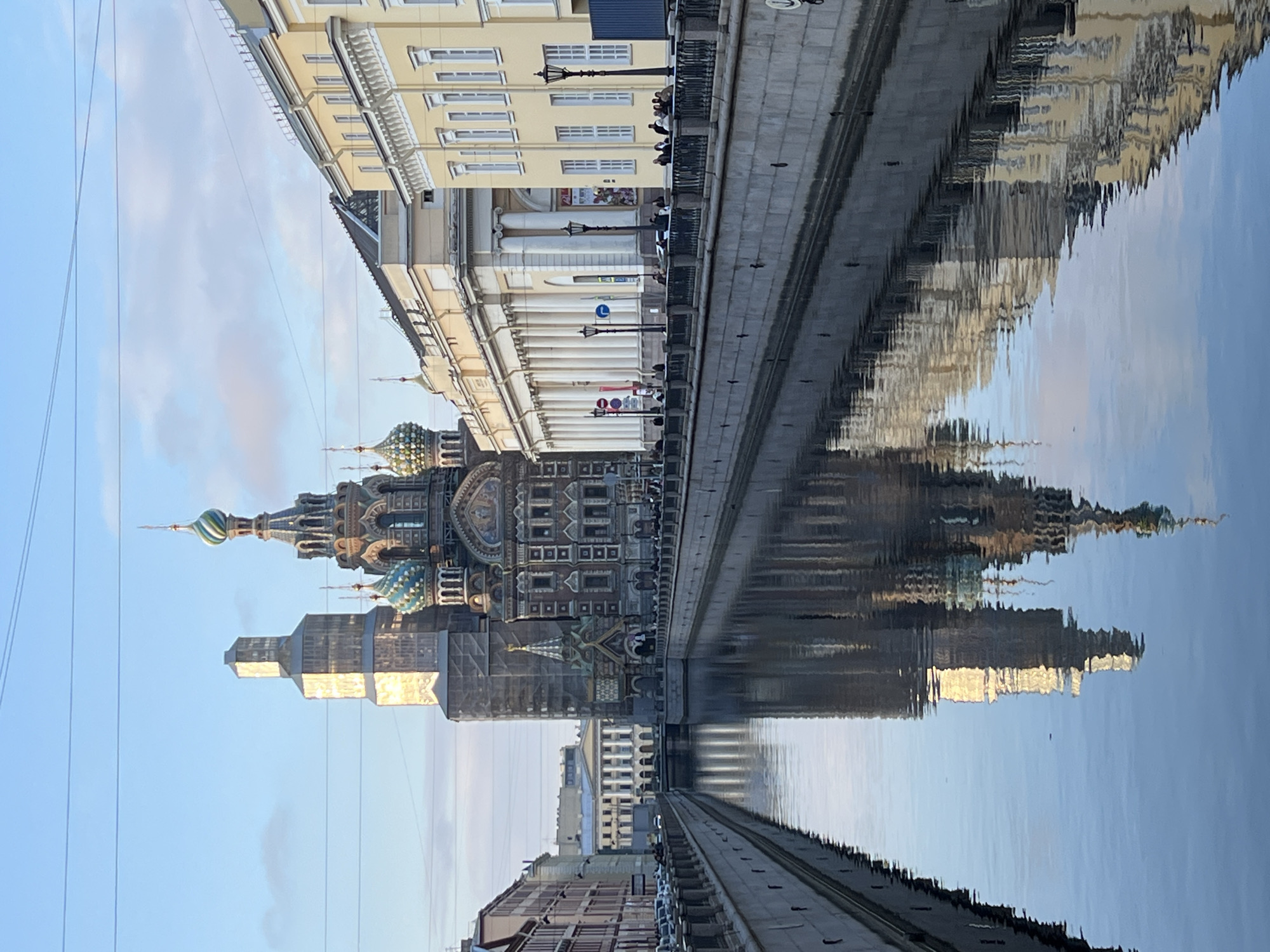 The image depicts the Church of the Savior on Spilled Blood in Saint Petersburg, Russia, reflected in the Griboyedov Canal. The canal bank features a stone embankment with people walking along it. Adjacent to the embankment, buildings are visible, including one with a colonnaded facade. The church's colorful domes and intricate facade are mirrored in the water. The sky is partly cloudy.