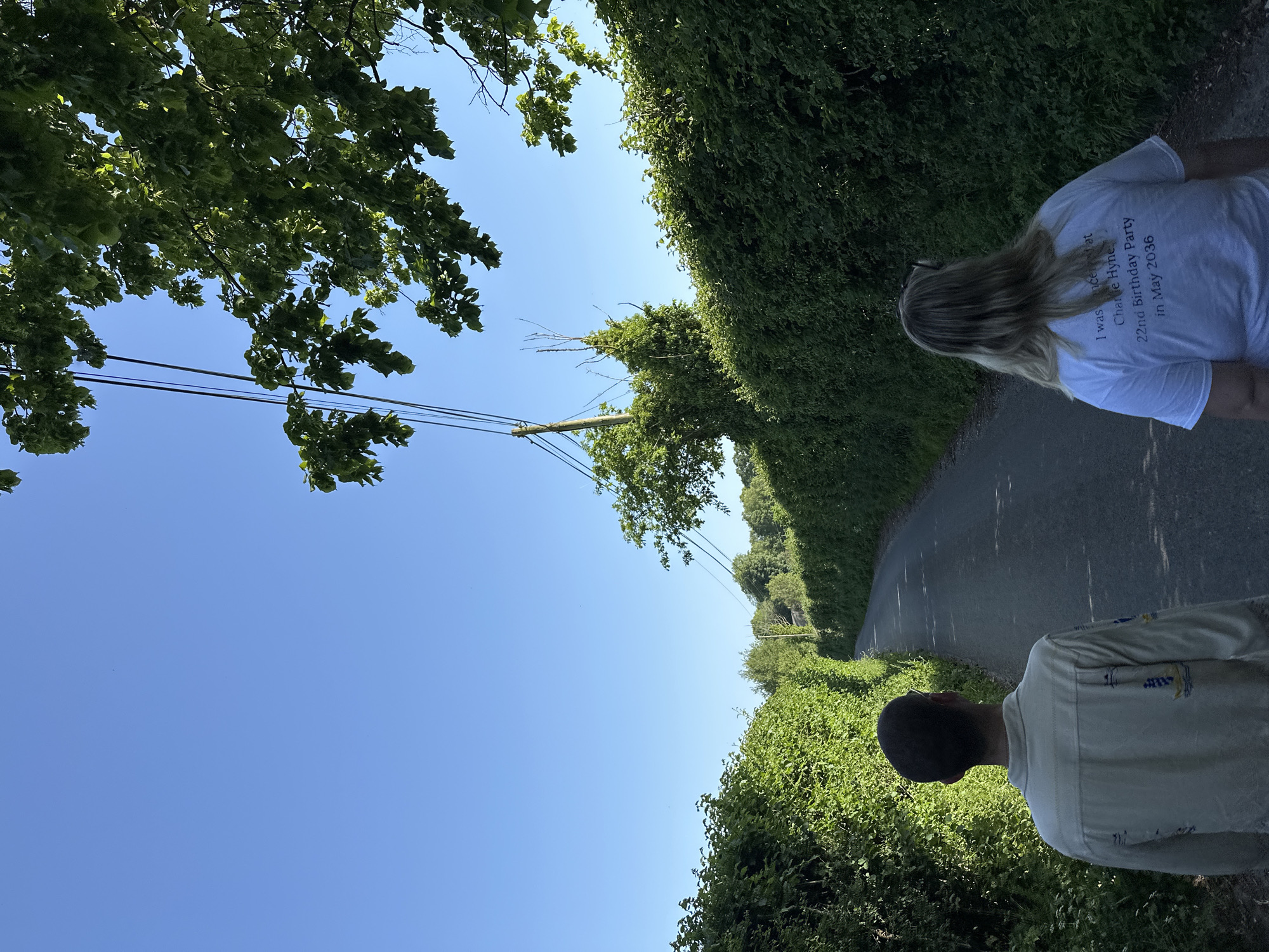 The image shows a paved road running between high green hedges under a clear blue sky. Power lines run along the left side of the road. Two people are partially visible. The head and shoulders of a person with dark hair are on the bottom right and the back of a person with long blond hair and a white shirt are on the top right. The shirt has text on the back that says "I was innocent at Charlie Hynes 22nd Birthday Party in May 2036." The location is unknown.