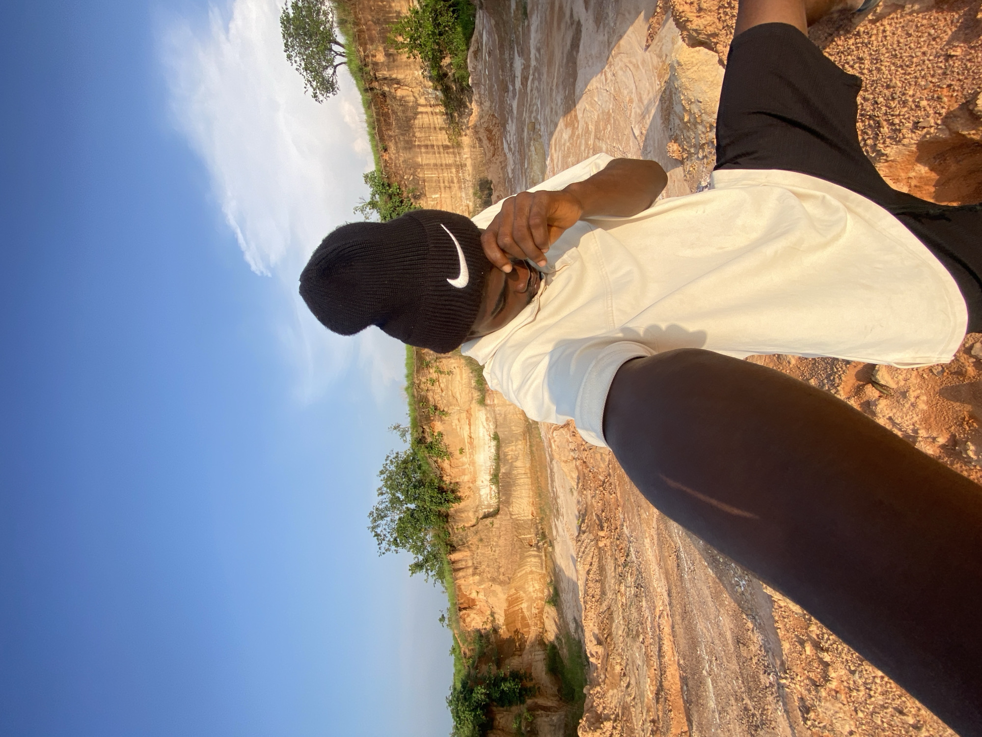 In Ufu Esioni, Nigeria, a person wearing a black Nike beanie and a cream-colored t-shirt stands in a quarry. The person is holding their right hand to their face. The ground is a mix of dirt and rocks, with layered sediment walls visible in the background. Green foliage grows on top of the quarry walls and in scattered patches around the site. The sky above is clear blue with a few wispy clouds. The sun casts a warm light on the scene, creating shadows.