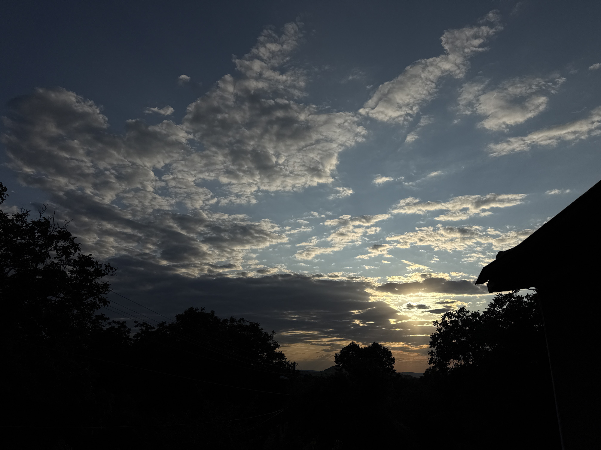 The image captures a silhouetted landscape at sunset, with a dramatic sky filled with a mixture of dark and light clouds. The sun's rays are breaking through the clouds, creating a warm glow on the horizon, suggesting the location is Krushevo, Bulgaria, known for its picturesque sunsets. In the foreground, the silhouettes of trees and a portion of a building are visible, creating a stark contrast against the vibrant sky. The overall scene evokes a sense of tranquility and natural beauty.
