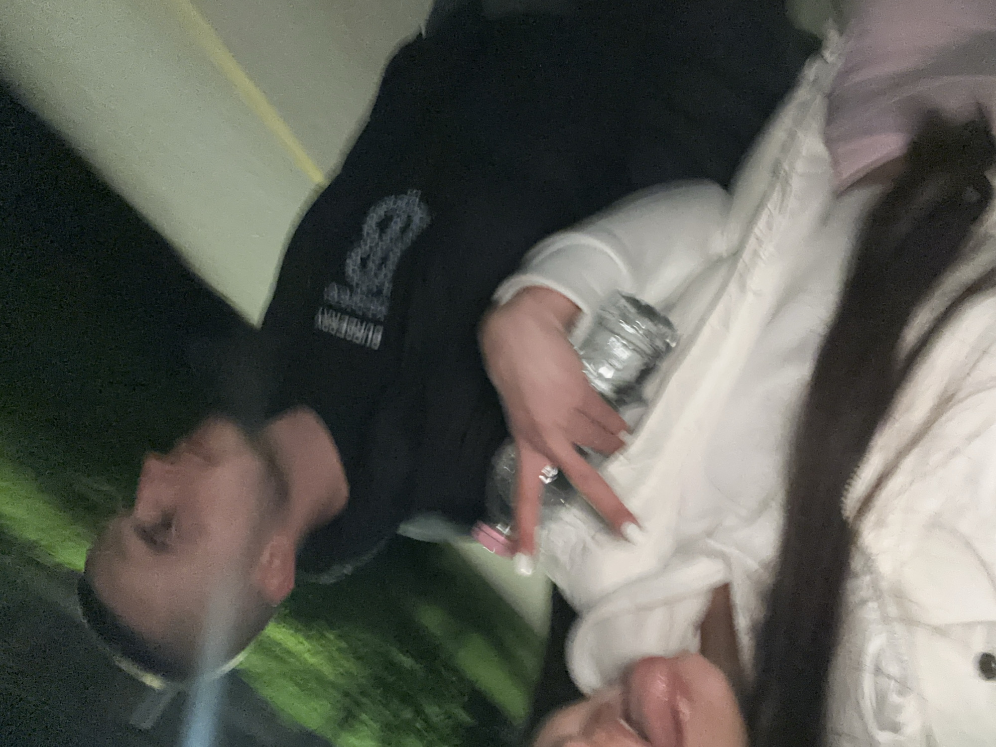 A blurry photograph shows two people. One person is a white male with short hair wearing a black sweatshirt with a "Burberry" logo. The other person, likely a female, is wearing a white jacket. Both are holding a water bottle. The location is not discernible. No specific details indicate the city of Plovdiv, Bulgaria. The environment appears to be nighttime.