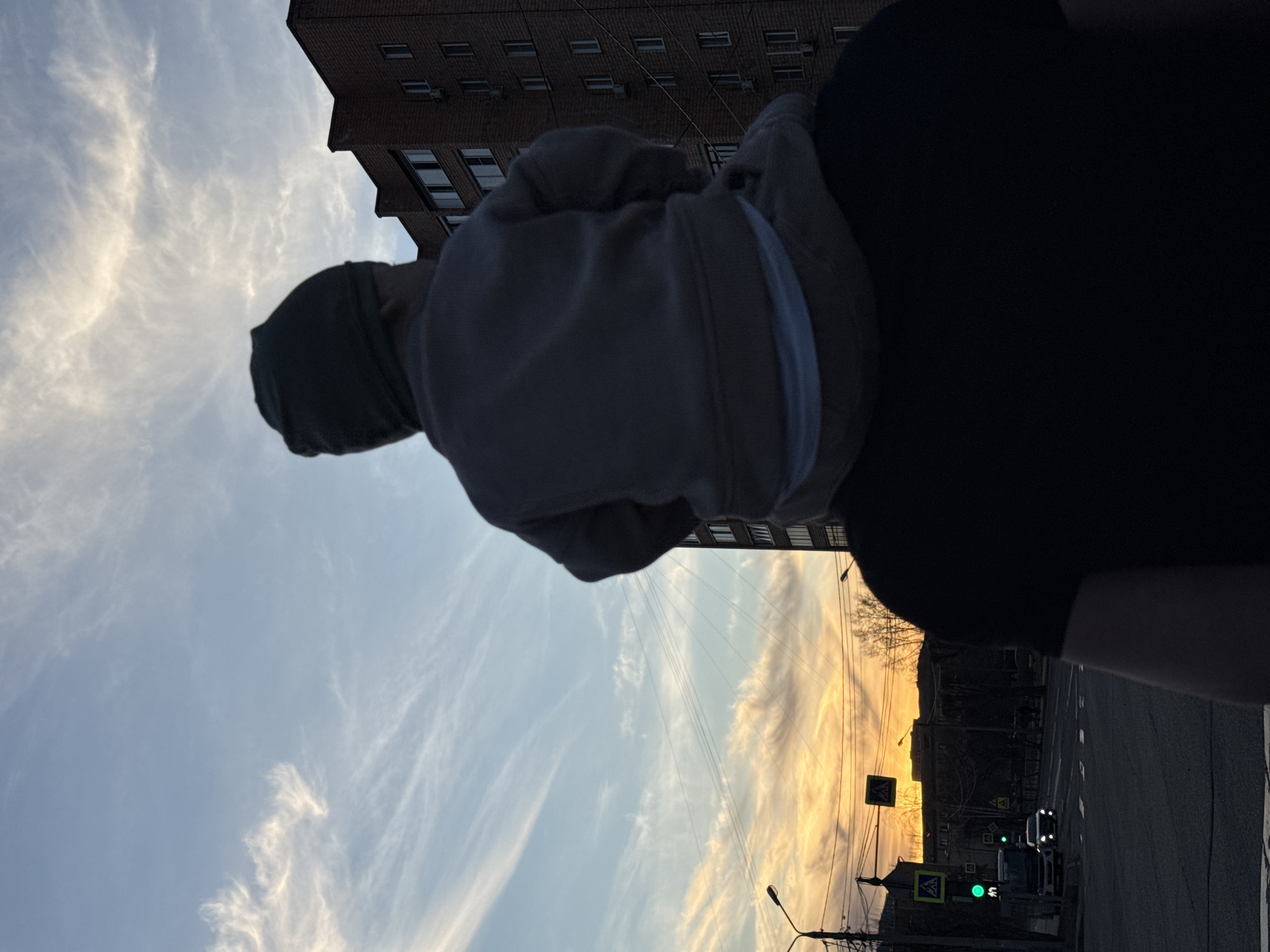 A low-angle, silhouetted image shows a person holding a small child on their shoulder, facing away from the camera. The child wears a hooded sweatshirt. In the background, there is a view of the city. The sky is visible with sunset clouds, and an apartment building and a street with traffic lights and cars are present. The location is Krasnoyarsk, Russia.