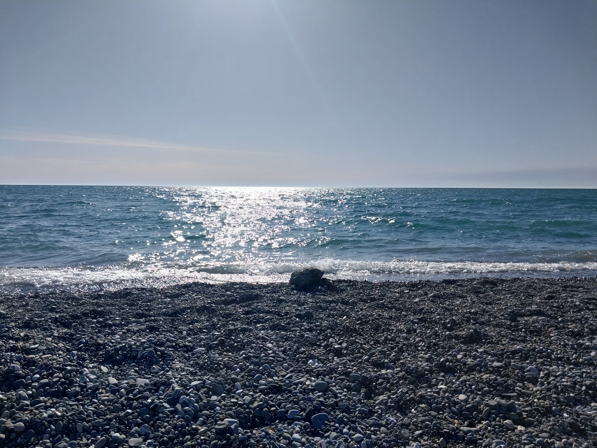 This image presents a serene coastal landscape, likely captured on a sunny day in Sochi, Russia. The scene is primarily composed of a pebble beach, the sea, and a vast sky, with no human subjects or man-made structures visible.
In the foreground, a wide expanse of dark, smooth pebbles covers the beach, showcasing a variety of grey and black tones. Towards the midground, the clear blue-green waters of the ocean meet the shore, with small, white-crested waves gently breaking. A notable feature on the pebbled beach, close to the waterline, is a single, moderately sized dark rock.
Beyond the immediate shoreline, the sea stretches out to the horizon under a bright, expansive sky. The sun, positioned high and slightly towards the left, casts a brilliant, shimmering path of light across the water's surface, creating a sparkling effect that extends from the horizon almost to the beach. The sky above is a pale, clear blue, with only faint, wispy clouds discernible near the distant horizon, indicating fine weather.
The overall atmosphere conveyed is one of tranquility and natural beauty. The time of day appears to be daytime, likely late morning or early afternoon, given the intensity and position of the sun. There is no visible text in the image.