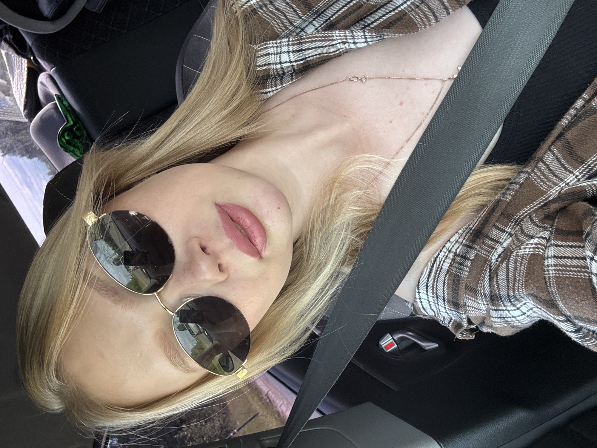 The media file is a close-up, high-angle, portrait-oriented photo of a young blonde woman inside a car, likely in Chaikovsky, Russia. She is lying down, with her head resting on a surface and her body angled upwards. She is wearing a brown and white plaid shirt, sunglasses with dark lenses, and a delicate rose gold necklace. A dark gray seatbelt crosses diagonally over her chest and shoulder, indicating she is the driver or a passenger. Her lips are painted a light pink. The background shows the car's interior, including a black car door handle with a red accent, and hints of the outside world reflected in her sunglasses, suggesting a bright, possibly sunny day. The overall mood is casual and relaxed.