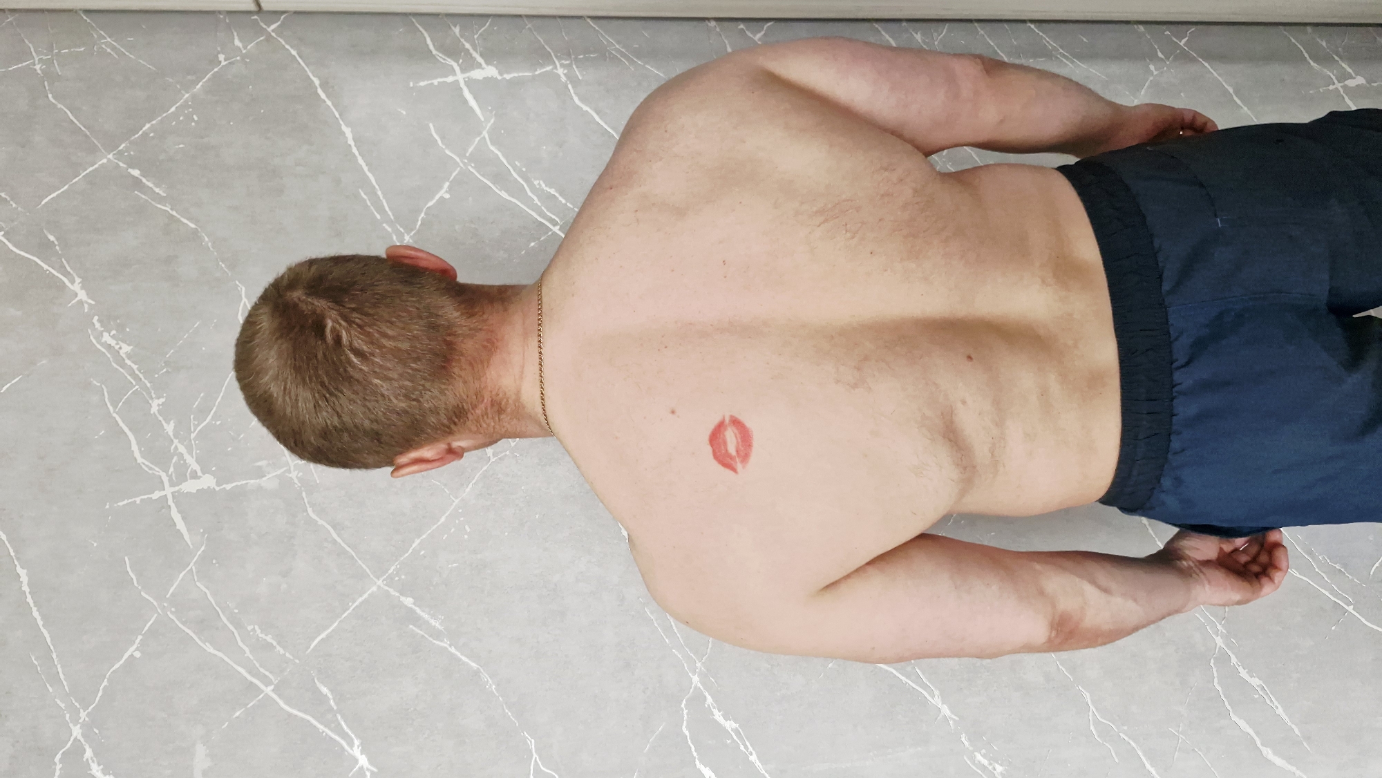 The image displays an adult male lying prone on a light grey floor with distinct white, marble-like vein patterns. He is shirtless, revealing his back and arms, and is wearing dark blue shorts with an elastic waistband. A thin golden chain is visible around his neck. A prominent red lipstick mark, clearly shaped like a pair of lips, is present on his upper back, slightly to the right of his spine. The man's head is turned to the left, showing the back of his head and his right ear. His arms are positioned along his sides, with his hands resting on the floor. The setting appears to be indoors, likely on a hard, tiled or polished concrete surface. The activity shown is simply the man lying down. No specific emotions are discernible, as his face is not visible, and there are no visible location cues within the image to suggest the provided context of Otchor, Russia. There is no visible text in the image.
