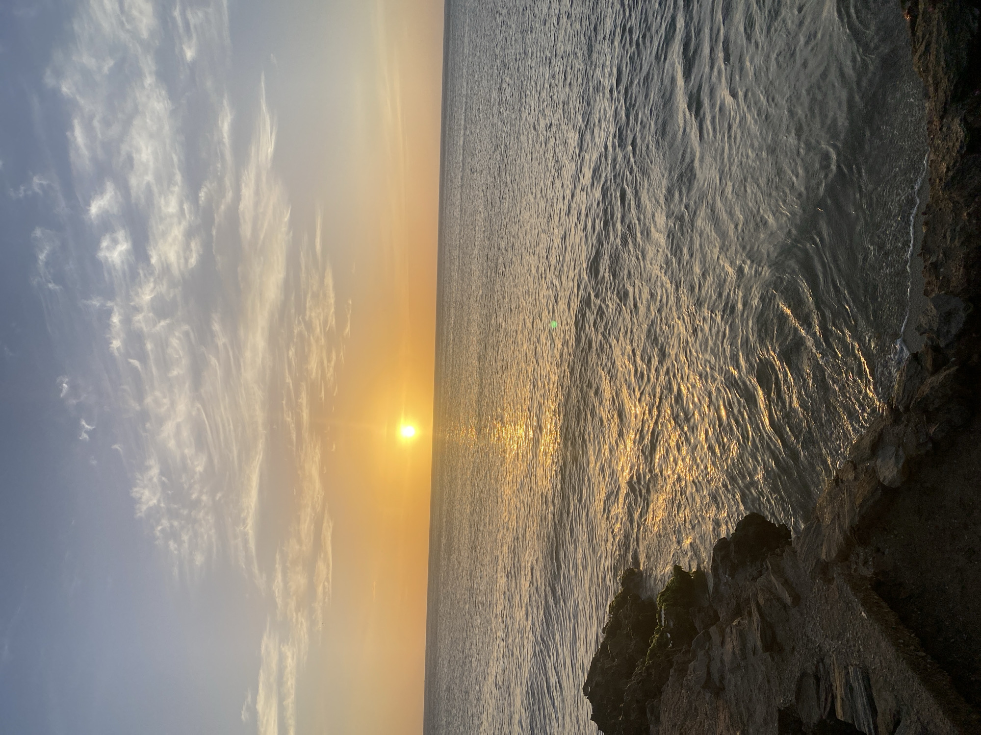 The image captures a serene sunset over the sea in Juan Griego, Venezuela. The sun, a bright orb of golden light, is positioned just above the horizon, casting a shimmering path across the gently rippling water. Wispy clouds, tinged with orange and yellow hues, stretch across the pale blue sky. In the foreground, rough, dark rocks form a rugged coastline, with some patches of green moss clinging to their surfaces. The scene evokes a sense of tranquility and natural beauty.