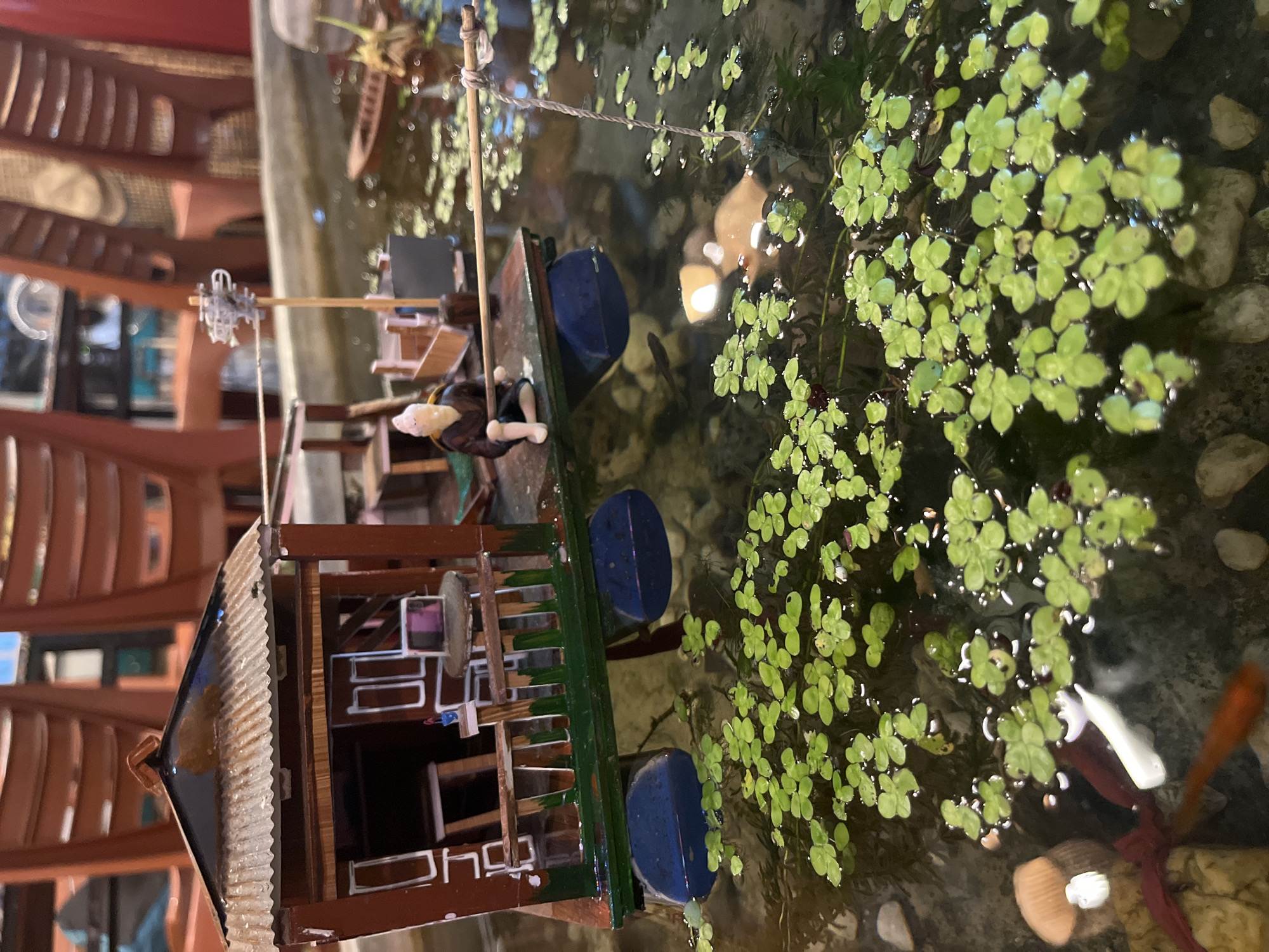 The image shows a miniature scene of a boat with a house on it, floating in a shallow body of water filled with green duckweed and pebbles. A small figurine of a person is seated on the boat, holding a fishing rod. The boat has blue pontoons and a red and green structure with a corrugated roof. In the background, out of focus, are rows of red chairs, suggesting an indoor setting possibly a restaurant or cafe. The context of Bac Nha Trang Ward, Vietnam, is not directly depicted in the visual elements of the image itself, but rather provided as external information.