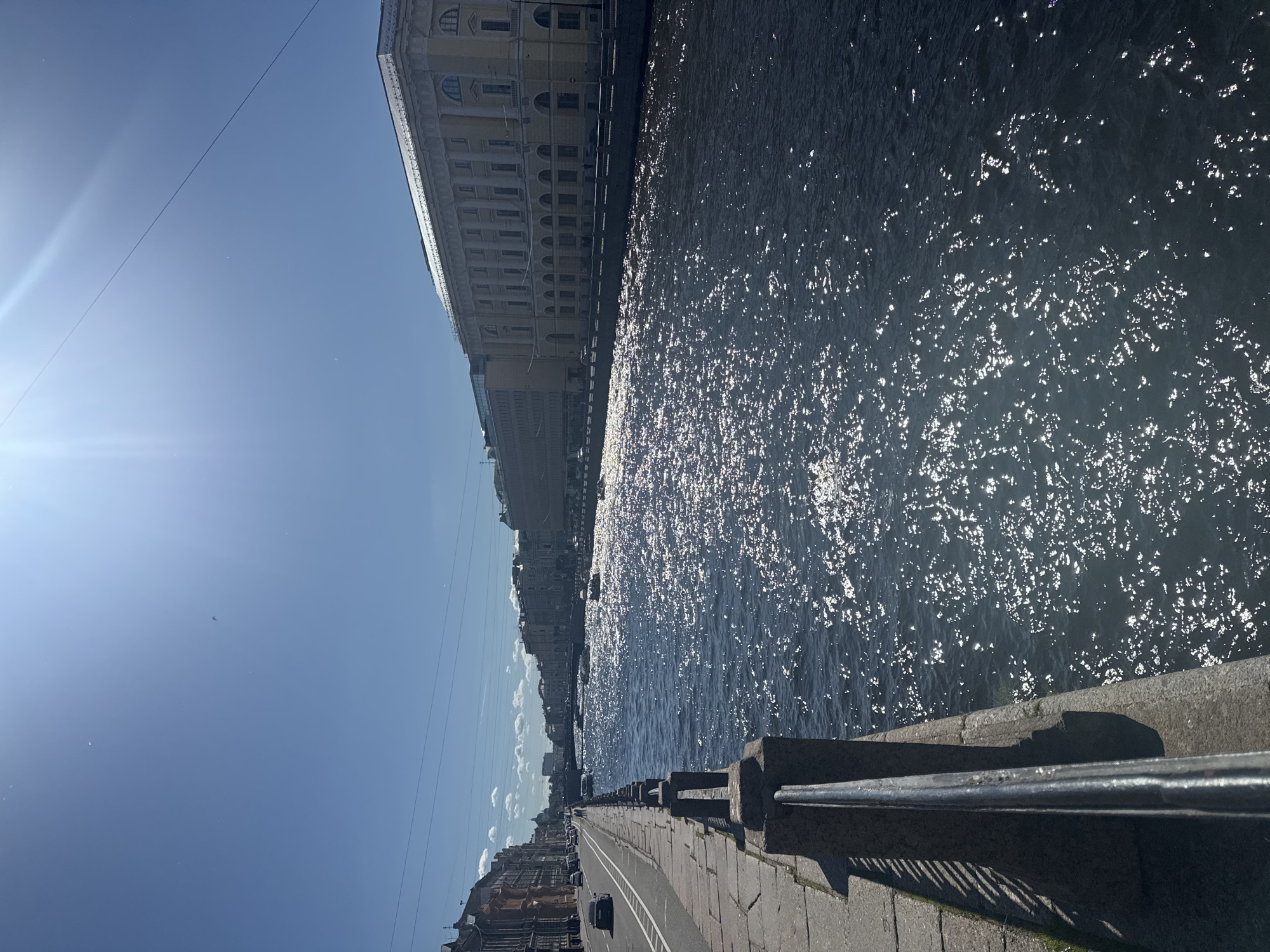 The image displays a sunlit scene along a river embankment in Saint Petersburg, Russia. The camera is positioned at an angle, looking down the embankment towards the horizon.
The primary subjects include a wide river reflecting the bright sunlight, creating a shimmering effect on its surface. On the left side of the river, a grand, multi-story building with classical architecture lines the embankment. Further down, more buildings and a street with parked cars are visible. The right side of the river features a stone embankment with a metal railing, casting long shadows on the paved walkway below. The sky is a clear, bright blue with a few wispy clouds.
The activity depicted is a tranquil daytime scene, likely a moment of leisure or transit. The bright sunlight suggests it is daytime, possibly late morning or early afternoon. The clear sky indicates fair weather. The architecture of the building and the presence of a wide river with stone embankments are characteristic of Saint Petersburg.
Notable details include the strong sun glare, the texture of the water's surface, the architectural style of the building, and the paved walkway with a railing. There is no visible text in the image. The overall mood conveyed is one of peacefulness and urban beauty.