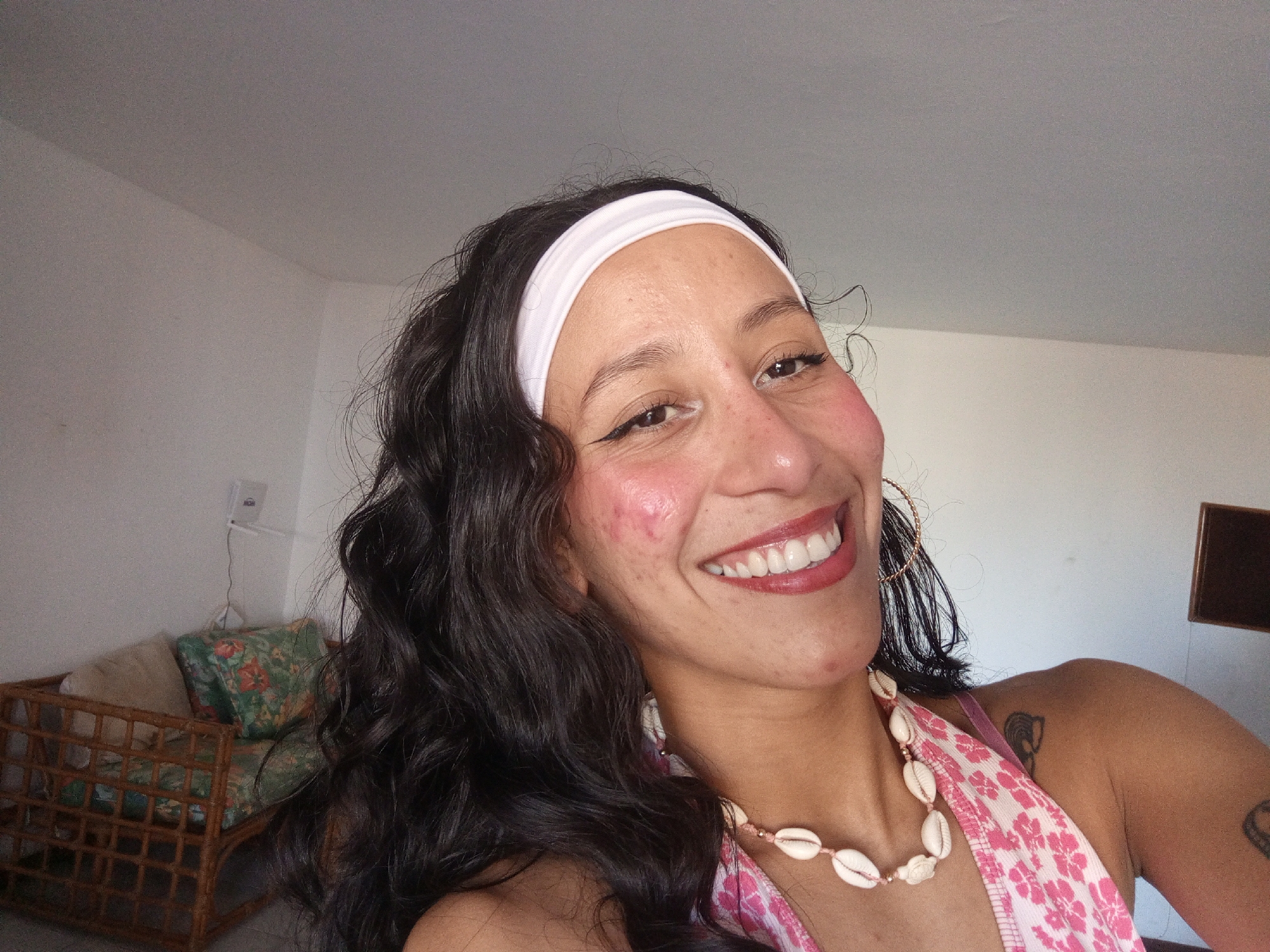 A woman with dark, wavy hair and a warm complexion smiles broadly into the camera, capturing a close-up selfie indoors in what appears to be a residence in Porlamar, Venezuela. She wears a white headband, a pink and white patterned halter-neck top, and a distinctive necklace made of white cowrie shells. A large gold hoop earring is visible on her right ear, and a small tattoo is seen on her left shoulder/upper arm. Her smile reveals white teeth, and her eyes are accentuated with eyeliner. Reddish blush is visible on her cheeks.
The background shows a simple interior with plain, light-colored walls and a ceiling. To the left, part of a wicker-style chair or sofa with green floral patterned cushions is visible, along with a white power outlet on the wall. The lighting suggests it is daytime. No visible text is present in the image. The overall mood conveyed is joyful and casual.
