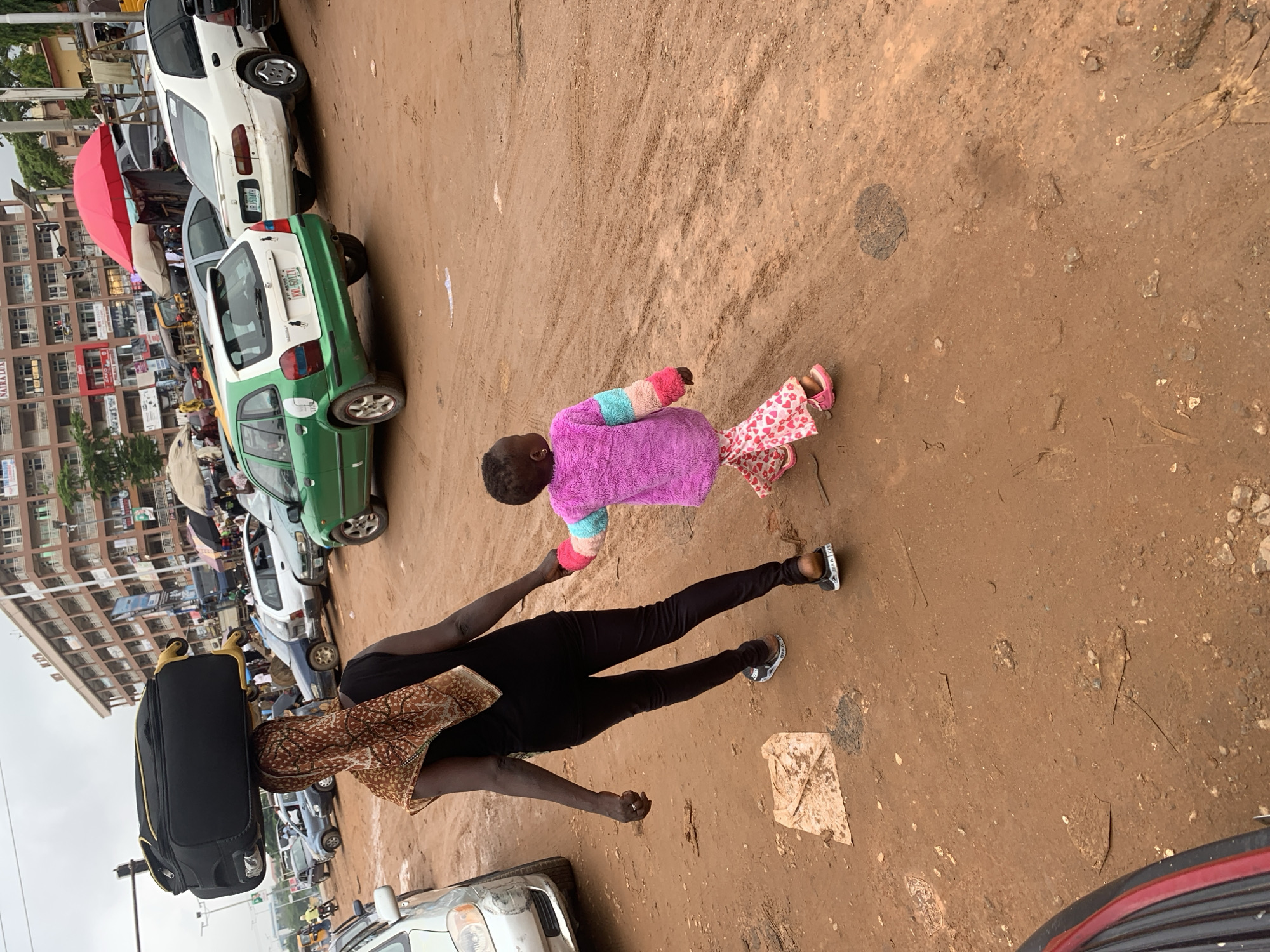 A woman with a suitcase balanced on her head walks away from the camera, holding the hand of a small child. The woman wears a black top and leggings, with a patterned headscarf. The child is dressed in a pink furry jacket with multi-colored sleeves and pink patterned pants, with pink sandals. They are walking on a dirt road in a busy urban area. In the background, there are parked cars, buildings, and market stalls. The overall scene suggests a bustling marketplace or transportation hub, likely in a city like Abuja, Nigeria, where such sights are common. The ground is dusty and uneven, with visible tire tracks and scattered debris. The sky is overcast, indicating a cloudy day.