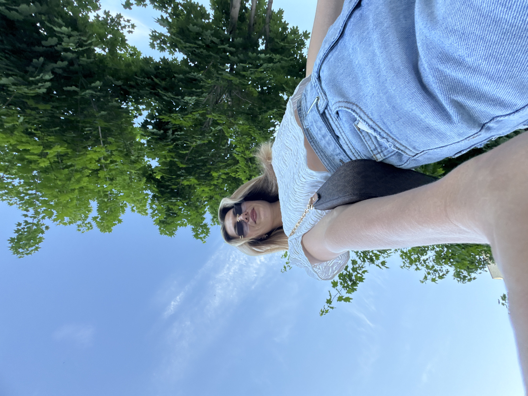 A low-angle, vertically-oriented shot reveals a person positioned in an unidentified urban location. The individual is wearing light-wash denim shorts and a white, textured top. They are accessorized with sunglasses. A brown purse is visible, held by a gold chain. Green trees and a blue sky with scattered clouds are in the backdrop.