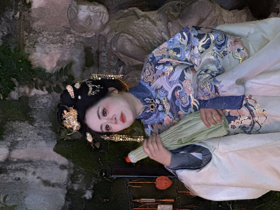 The image captures a female individual in traditional Chinese attire, positioned slightly left of center, in Yuzhong District, China. The setting is consistent with a cultural tourist area, specifically suggested by the visible "巴渝民俗风" (Bayu Folk Customs) inscription on a background wall, indicative of locations like the Hongyadong Cultural Tourist Area.
The subject is depicted from the chest up, facing right with her gaze directed slightly upwards. She wears an elaborate hairstyle featuring a large black bun adorned with gold-colored ornate hairpins, a crown-like piece, pearls, and dangling chains. Her facial makeup includes prominent rosy blush on her cheeks, defined eyebrows, and shimmery eyeshadow. She is dressed in a richly embroidered blue top featuring large white crane motifs, clouds, and floral patterns in blue, pink, and gold, with a high collar embellished with black and gold details. Over this, a flowing, light-colored (possibly off-white or very pale green) outer robe is draped. Her hands hold a folded, light green umbrella, distinguished by a red tip at its peak.
The background consists of a dark, textured stone or concrete wall with minimal vine or moss growth. To the left, a black pole or railing features small, square-shaped charms or plaques, some with red heart designs, hanging by chains. To the right of the subject, a weathered stone statue of a young boy or child is partially visible. The aforementioned "巴渝民俗风" characters are faintly visible on the wall behind this statue. The scene is illuminated by artificial light, creating a focused glow on the subject and localized shadows in the immediate environment.