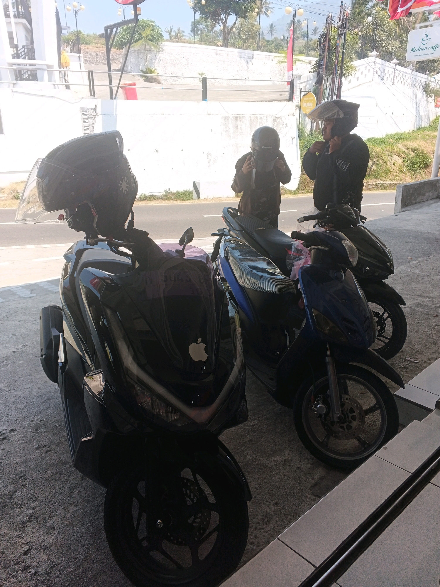 In Pakopen, Indonesia, two individuals stand near parked scooters. The person on the right, wearing a black jacket and helmet, is adjusting their helmet strap. To their left, another person, also in dark attire and a helmet, appears to be speaking or looking towards the first individual. Three scooters are visible: a black one in the foreground with an apple logo on its front, a blue one to its right, and another dark scooter behind the blue one. The scene is outdoors, with a paved area and a road in the background, suggesting a roadside location. Buildings with white walls and decorative elements are visible beyond the road, and trees and sky complete the environment.