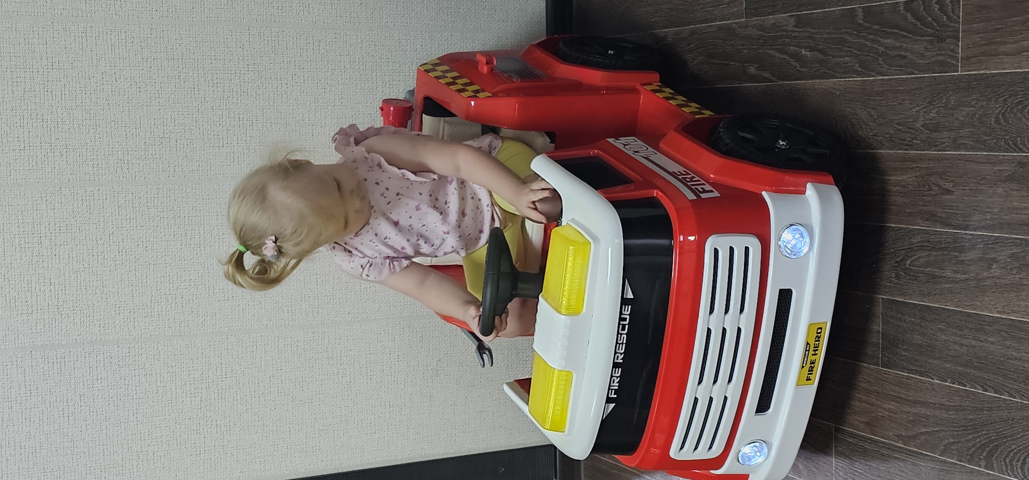 A young child with blonde pigtails, adorned with a small pink bow in her hair, is seated in a red toy fire truck. She wears a pink shirt with a floral pattern and yellow pants. The child is actively engaged with the toy, her hands on the steering wheel. The fire truck is bright red with white accents, featuring yellow headlights and a grill. Text visible on the truck includes "FIRE RESCUE" and "FIRE HERO." The scene appears to be indoors, with a textured light-colored wall on the left and dark wooden flooring on the right. The lighting suggests it could be daytime.
