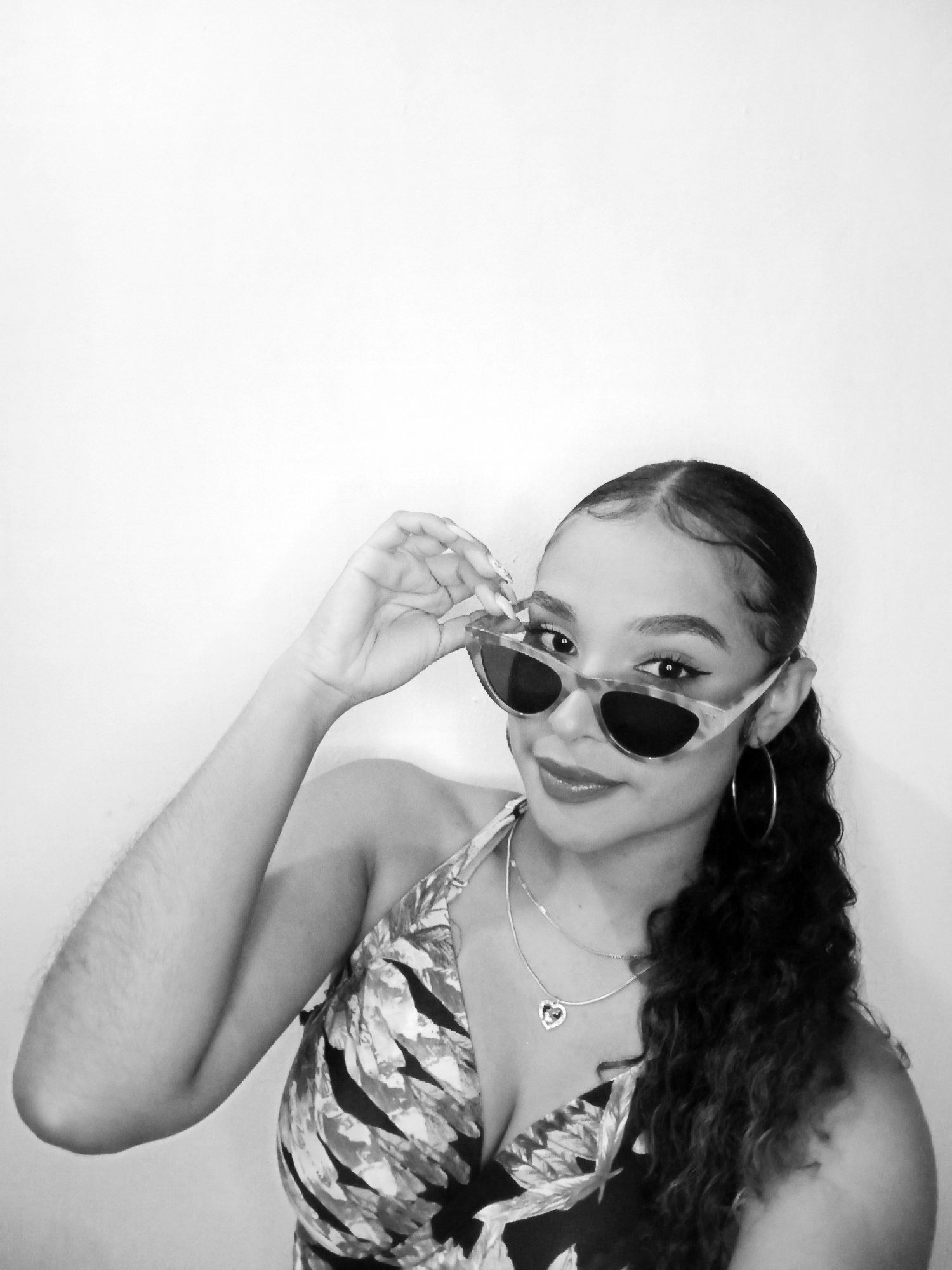 A monochrome portrait of a young woman holding sunglasses. She has dark hair, styled with defined waves and a ponytail. She wears a floral print top and silver jewelry, including a heart-shaped pendant necklace and hoop earrings. The background is a plain white wall. The location of the image in Juan Griego, Venezuela, is not determinable from the image alone.
