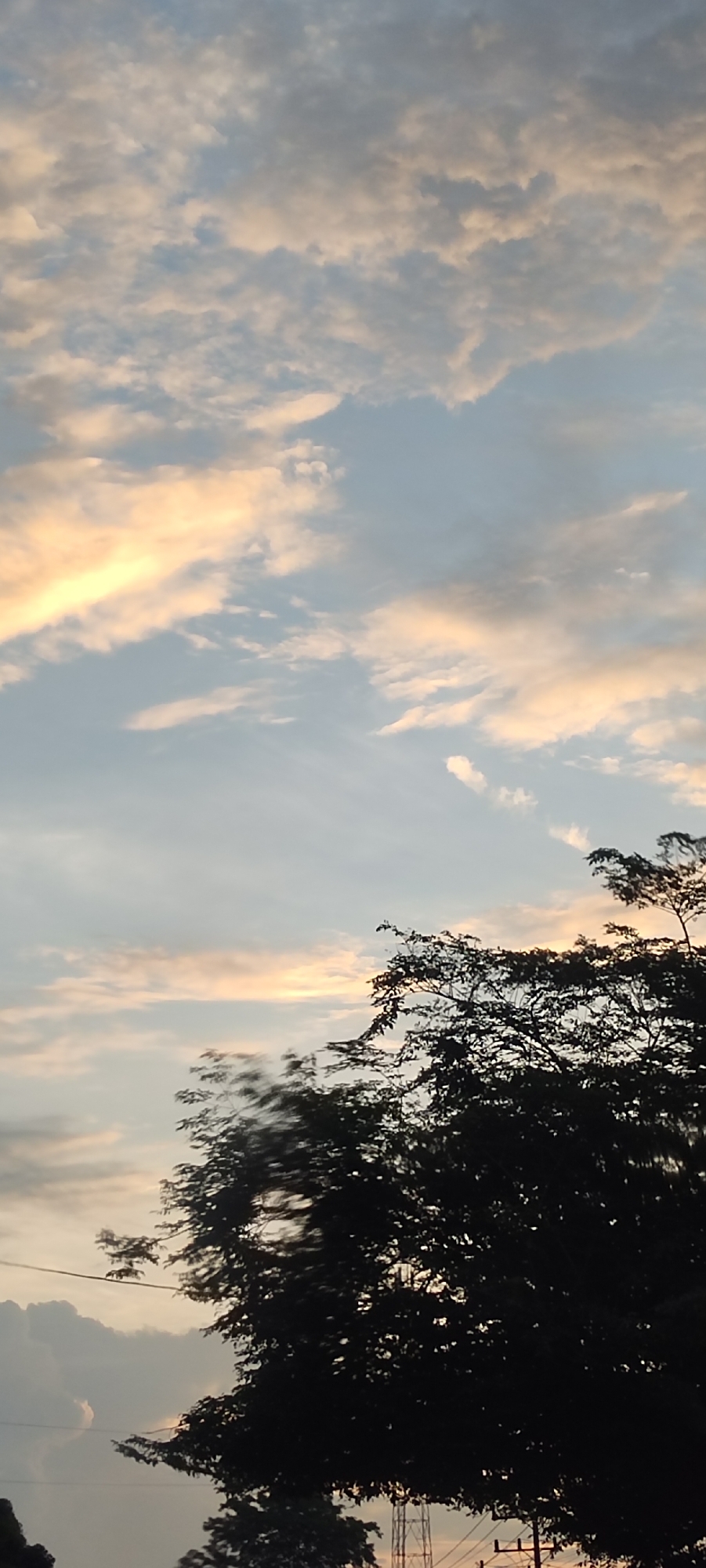 The image is taken outdoors in Aluegunto, Indonesia, depicting a partly cloudy sky at sunset or sunrise. The clouds are a mix of white, gray, and light orange hues, indicating the presence of sunlight. In the lower portion of the image, the silhouette of a tree with dense foliage is visible. Wires and an antenna structure can be discerned among the trees.