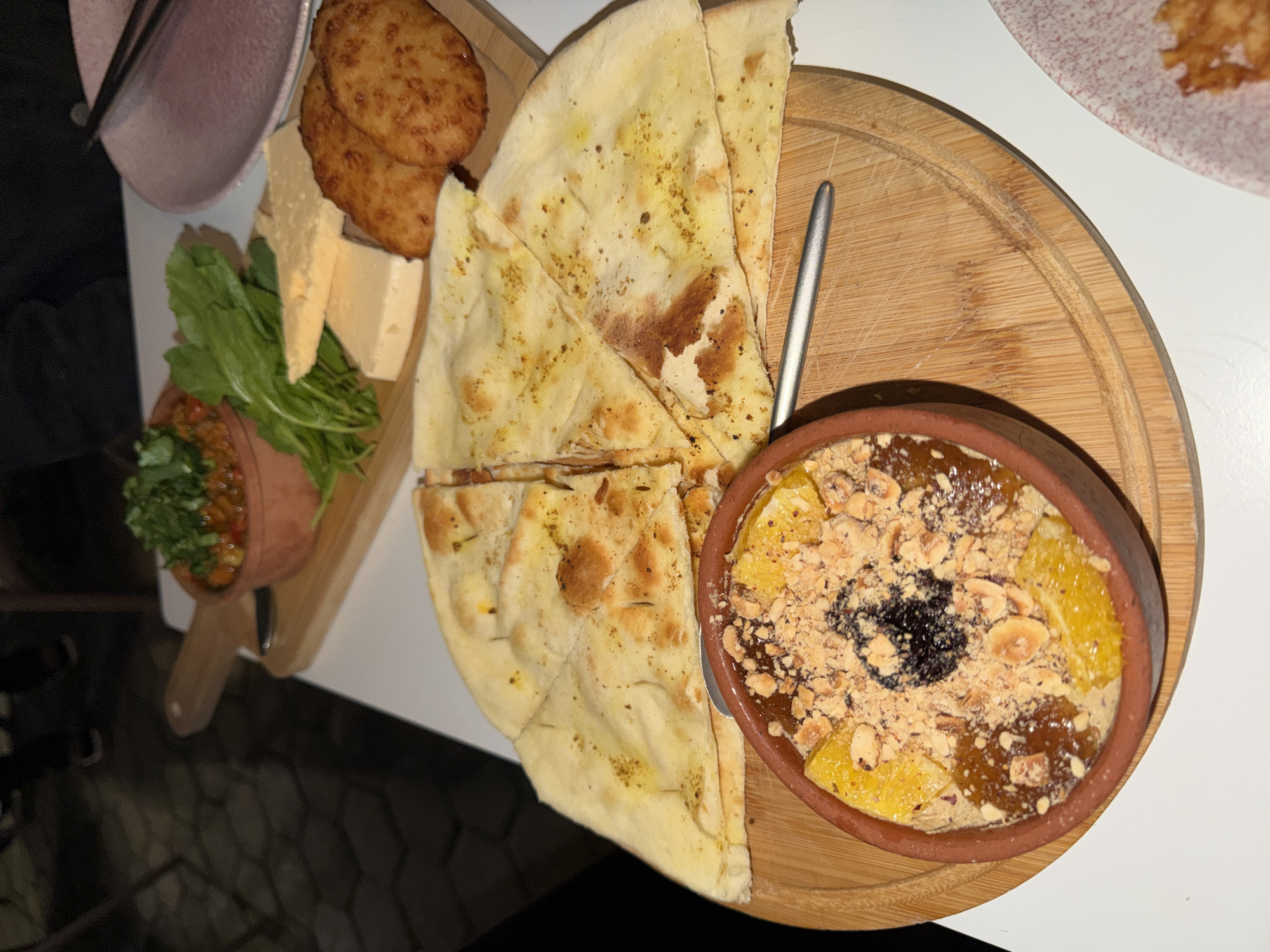 On a table, a round wooden board holds flatbread, a small pot of dessert topped with nuts and fruit, and a small metal spoon. A wooden cutting board next to it holds cheese, arugula, and a small pot with a vegetable mix. Behind the cutting board is a plate with more food. The location may be Batumi, Georgia.