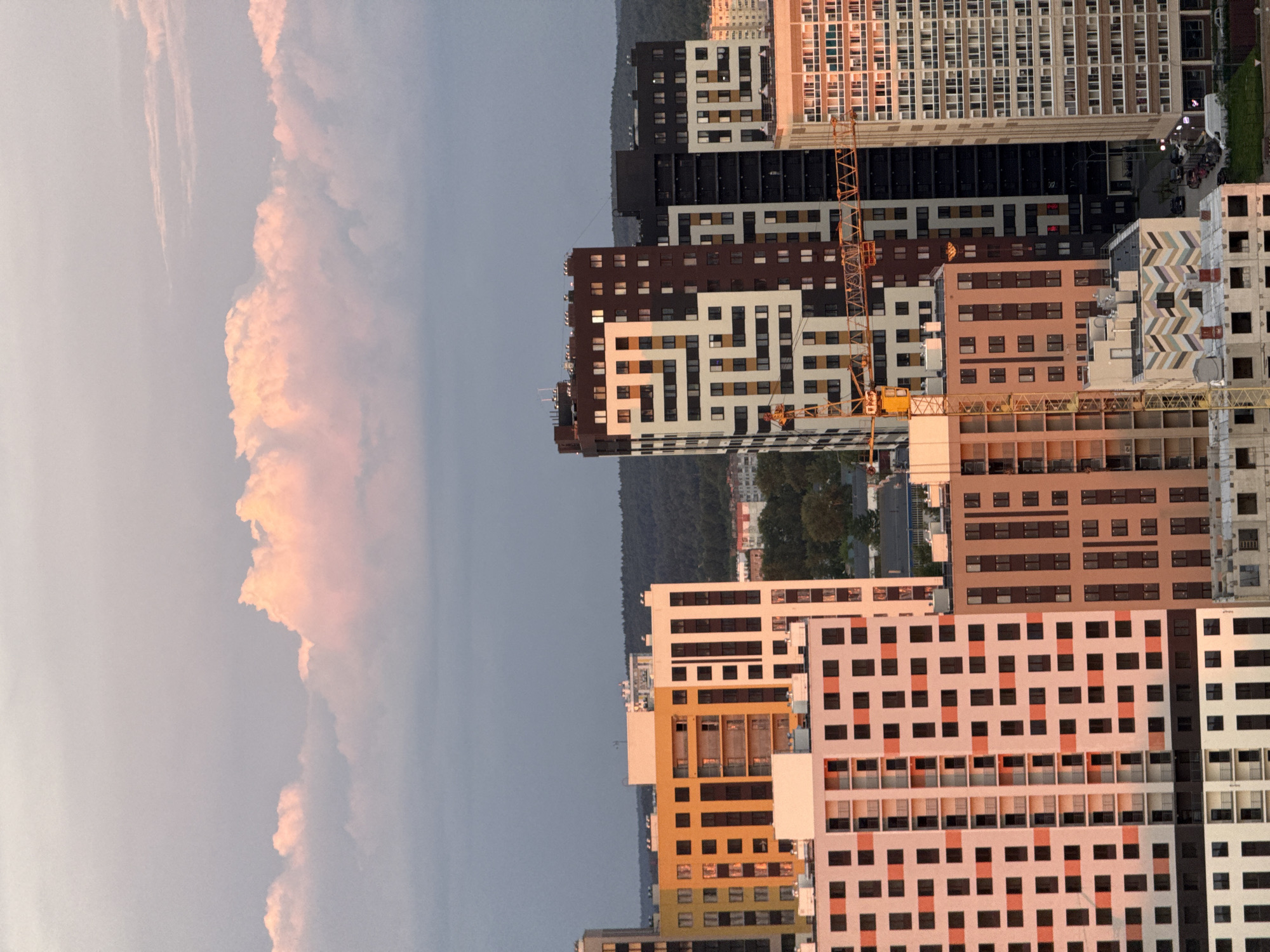 The image captures a modern urban landscape in Sokol, Russia, seen from an elevated perspective. The scene is illuminated by the warm, low-angle light of either sunrise or sunset, creating a golden hour effect.
In the upper and left parts of the frame, a pale blue-grey sky dominates, featuring large, voluminous clouds with striking peachy-pink and orange hues, catching the warm sunlight. Below the clouds and in the background, a dark green band of trees, likely a forest, stretches horizontally across the horizon.
The majority of the image, particularly the right and lower sections, is filled with numerous high-rise residential buildings. These modern structures display a variety of architectural styles and colors, including facades with dark brown, white, beige, orange, and reddish-brown panels. Many buildings exhibit a grid-like pattern of windows, some of which reflect the golden light. The presence of at least two prominent yellow tower cranes indicates active construction. The buildings appear to be either newly completed or still under development, suggesting a rapidly expanding residential area. No discernible text is visible.