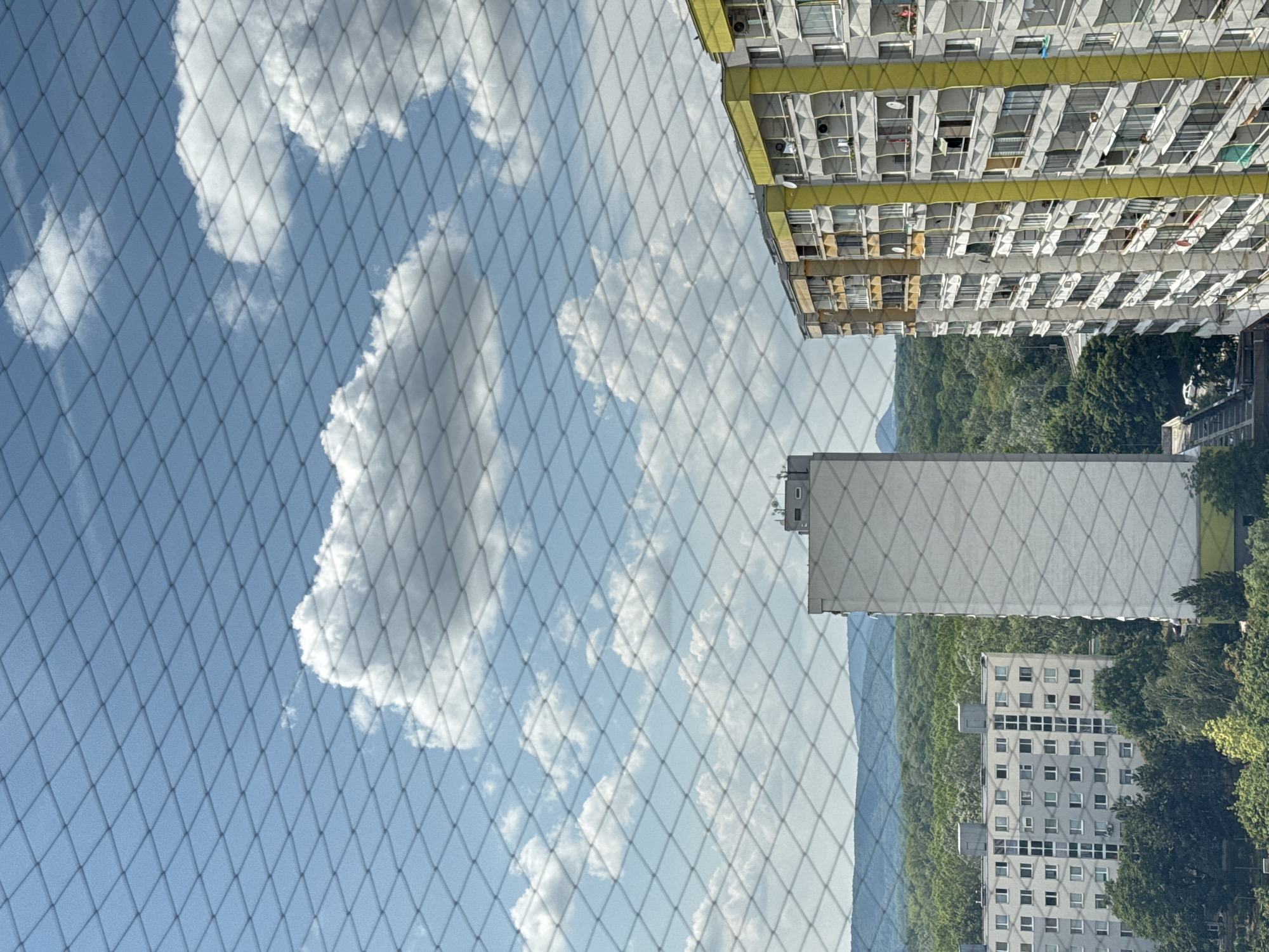 The image displays a view of residential buildings in Bratislava, Slovakia, seen through a diamond-patterned net. In the foreground, the net obscures parts of the scene. In the middle ground, there are several multi-story apartment buildings. One building on the right is tall and gray with yellow accents and numerous balconies. To its left, another apartment building is visible, also with balconies and windows. Further to the left, a more distant, slender, gray high-rise building stands out. The background features a dense forest with rolling hills and mountains in the distance under a blue sky with scattered white clouds. The overall impression is an urban residential area bordered by natural landscape.