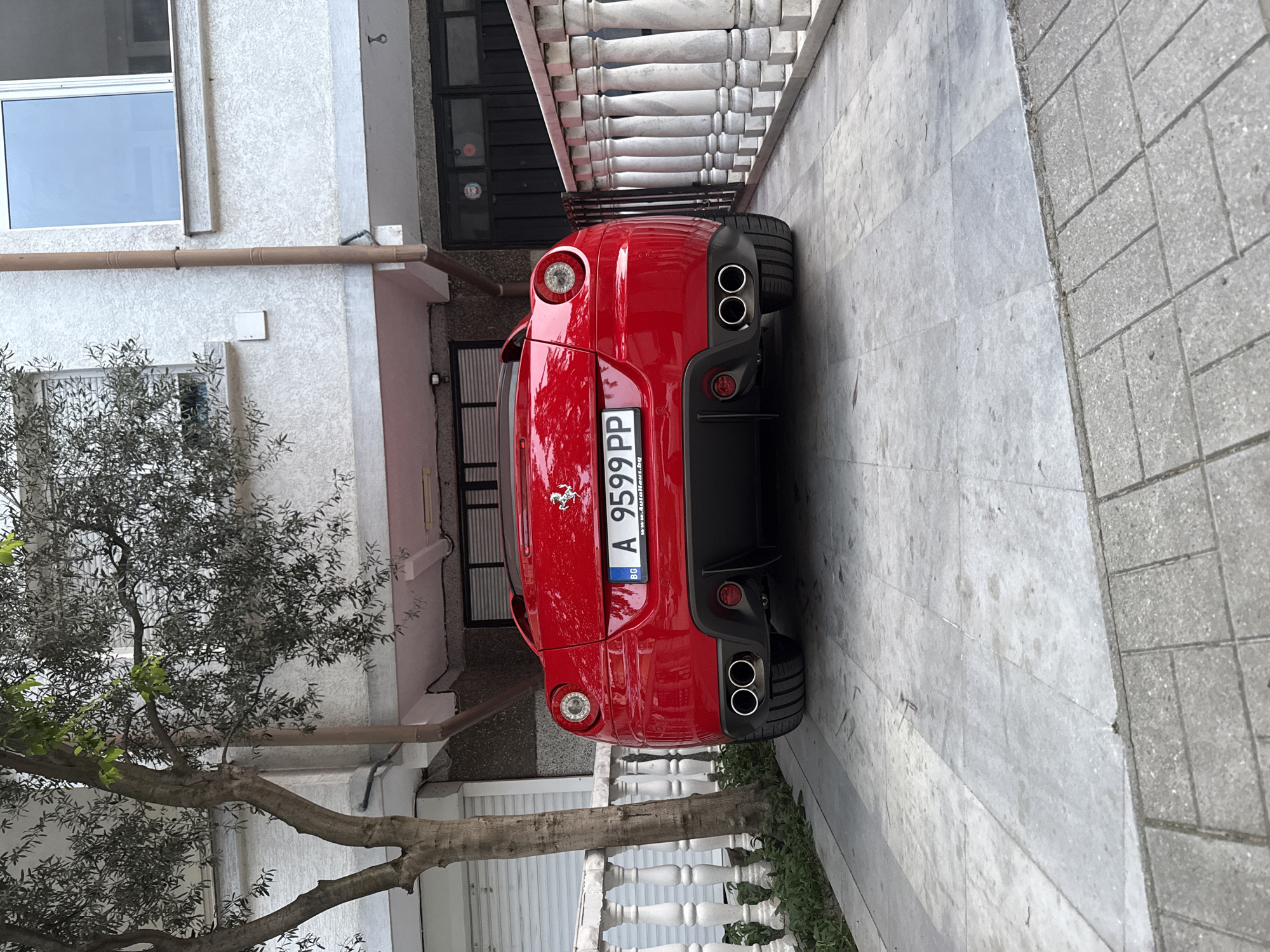 The image captures the rear view of a red Ferrari car parked in what appears to be a residential or semi-urban setting. The car's license plate is visible, reading "A 9599 PP", with the country code "BG" (Bulgaria). The car is positioned between a building and what seems to be a paved pathway.
The building features a combination of white stucco walls, metal railings, and a window on the upper level. A tree with dense foliage is visible on the left side of the frame, partially obscuring the building.
The pavement is made of large, light-colored tiles. The overall environment suggests a well-maintained or affluent neighborhood. The specific location within the city of Unknown, Unknown cannot be determined.