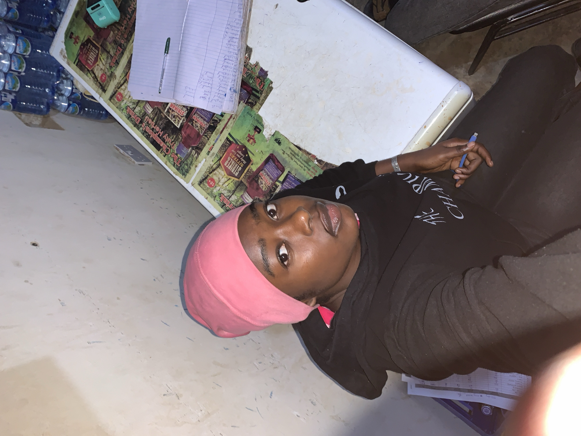 The image shows a person with dark skin wearing a pink head covering and a black hoodie. The person is seated with what appears to be a white plastic table in front of them. On the table are water bottles, a notebook with writing, a pen, and several promotional stickers or advertisements featuring a drink called "Striker Bitters." Some of the stickers are cut up. The table has a green and maroon advertisement attached to the top of it. The person is holding a blue pen in their hand. The background includes a cement wall and some papers stacked under the person's leg.
Based on the OCR text on the advertisements, the address "50. Iyesi Road, Ota Ogun Sta" is visible. Given that the OCR also says "The Champion's Drink," and based on the other text, the location could possibly be in the vicinity of the New Stadium, Nigeria.