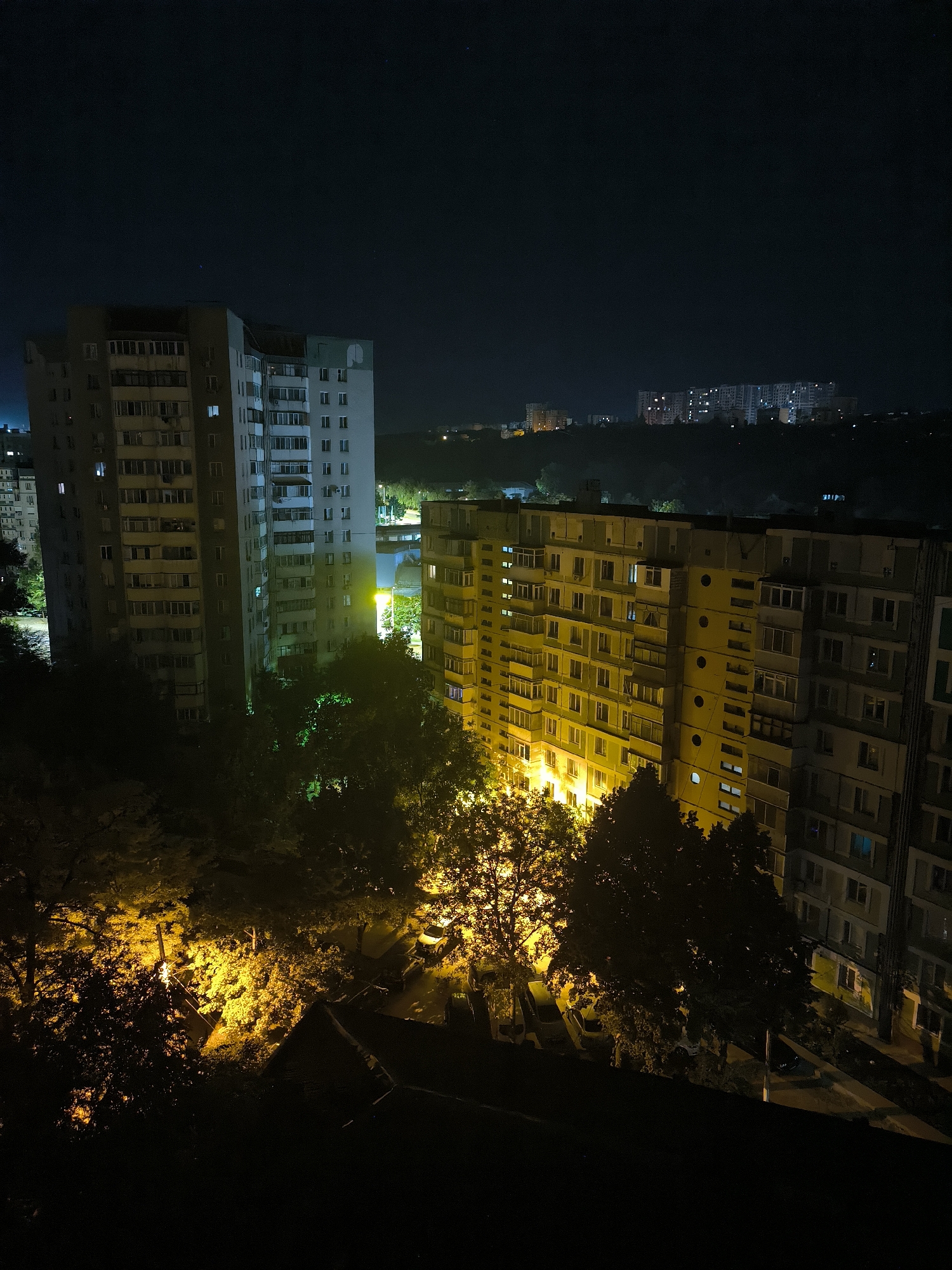 This is a nighttime, high-angle, long-shot photograph of an urban landscape in Chișinău, Moldova. In the foreground, silhouetted trees obscure the lower portion of the scene. To the left, a tall, multi-story apartment building with numerous windows, some illuminated, is visible. To the right of this building, another, wider apartment building is lit with warm yellow light emanating from many of its windows. Trees with illuminated foliage are situated between these buildings. In the middle ground, a street with parked cars is illuminated by streetlights. In the background, more apartment buildings are visible, some with illuminated windows, against a dark night sky with faint stars. The overall scene depicts a residential area at night.