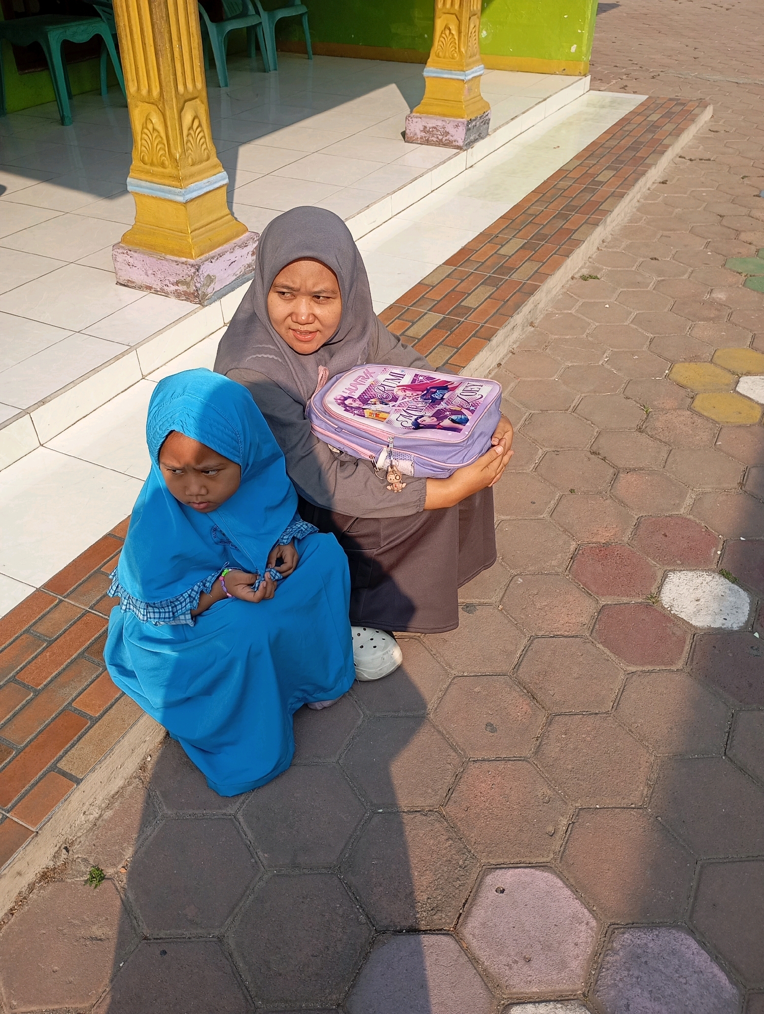 In a sunlit outdoor area in Babelan, Indonesia, a young girl in a bright blue hijab and dress sits on the ground, looking down with a pensive expression. Beside her, an adult wearing a grey hijab and a dark grey dress holds a light purple lunchbox adorned with cartoon characters. The lunchbox appears to have a zipper and a small charm attached. They are positioned on a textured sidewalk composed of hexagonal and rectangular paving stones. In the background, the base of a decorative column painted yellow and blue is visible, along with a tiled step leading up to a green wall and white tiled flooring. The scene suggests a moment of rest or waiting, possibly before or after attending school or an event.