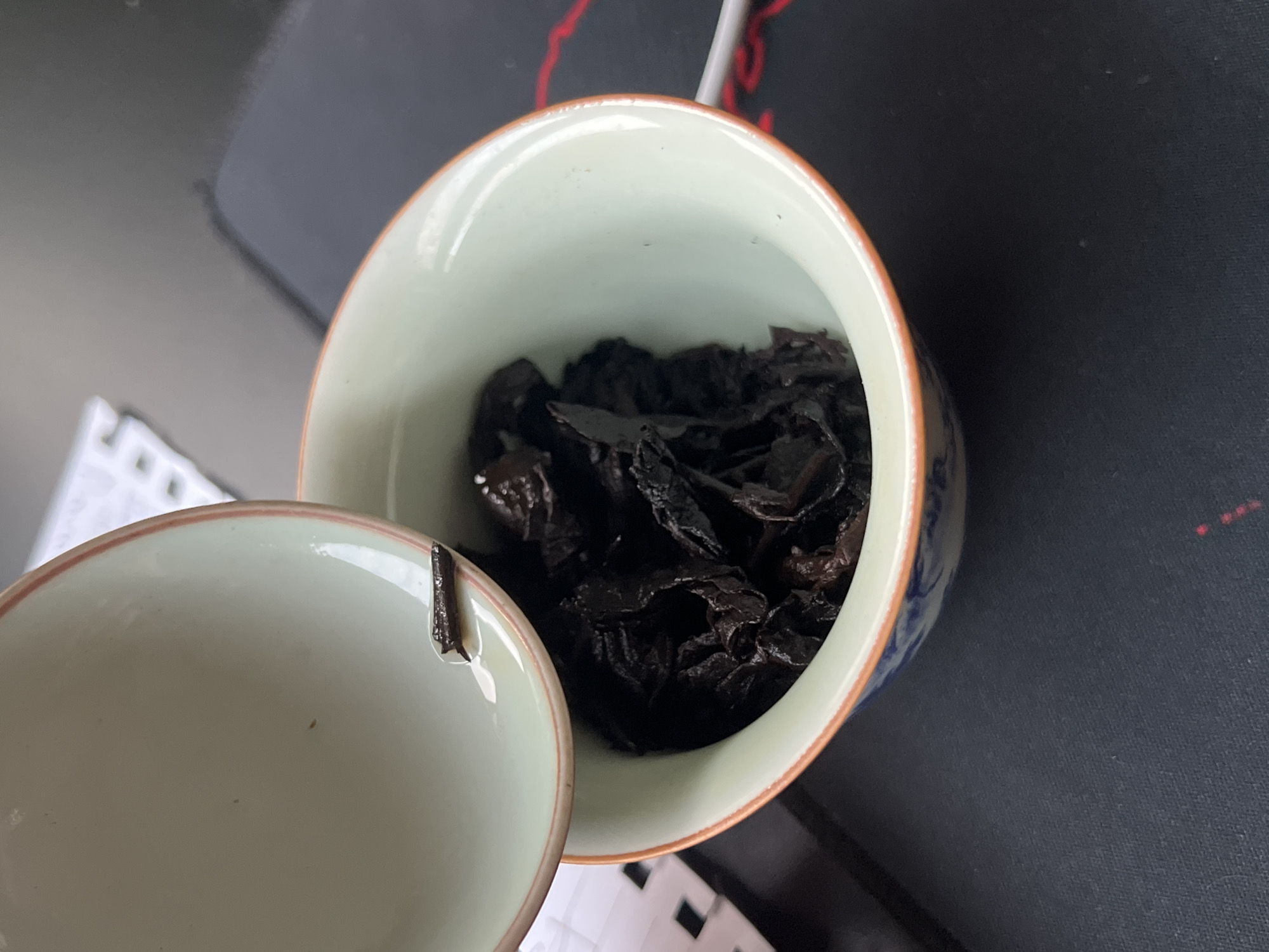 A high-angle shot depicts a light green and blue ceramic cup almost filled with dark, crumpled material. The cup is open, with a matching lid resting slightly askew next to it. A similar fragment of dark material is visible on the lid. The background consists of sections of a white keyboard, a black surface (possibly a mousepad), and a hint of a red cable.